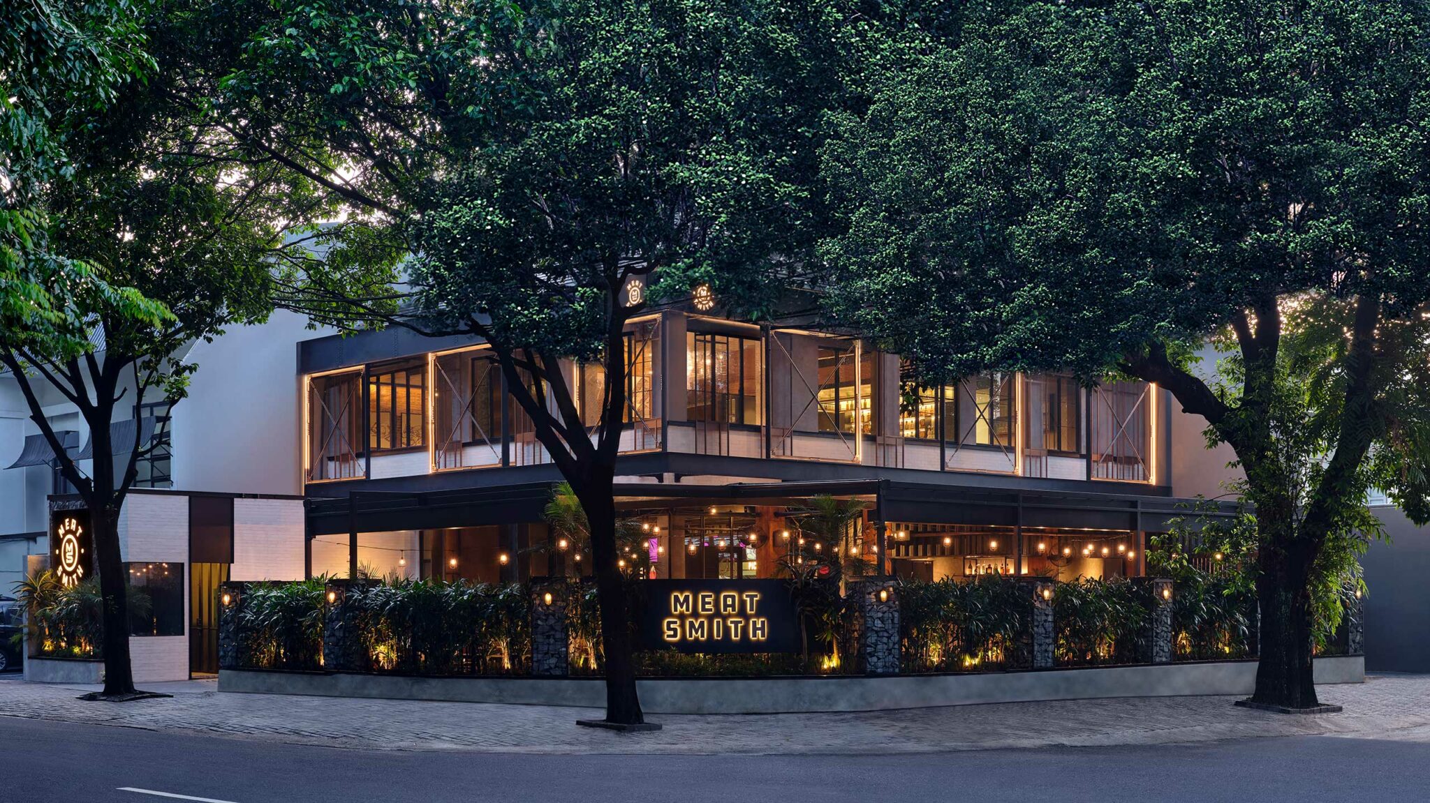 Exterior view of Meatsmith Jakarta, a two-storey contemporary grill restaurant designed with industrial steel framing, warm lighting, and lush landscaping.