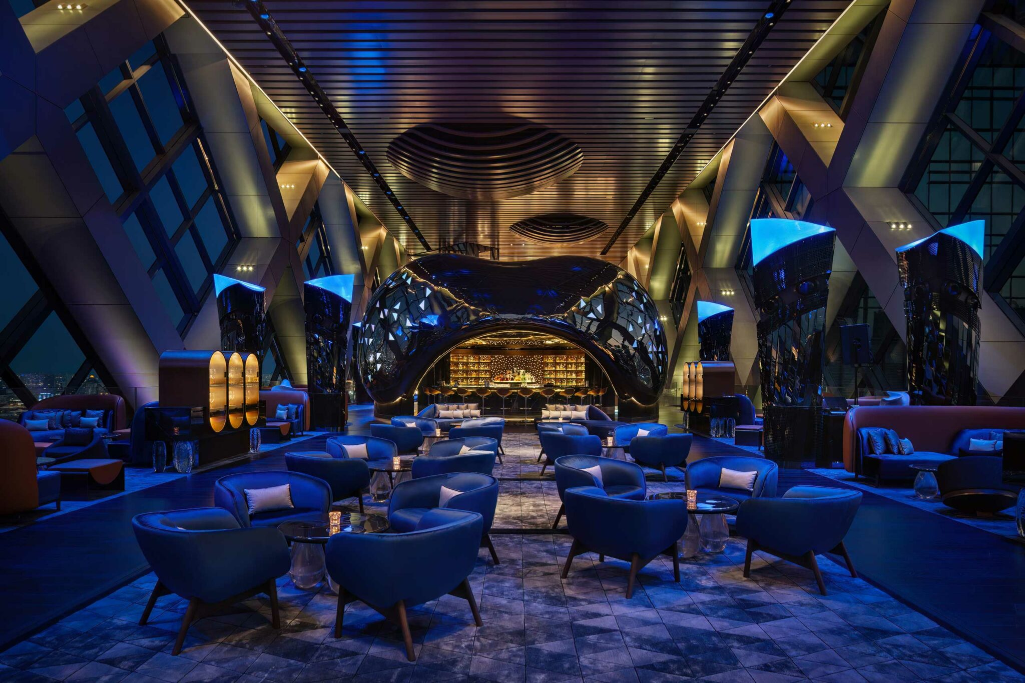 Full frontal view of Sphere’s sculptural pod enveloping the central bar, with mirrored black surfaces and triangular LED details, surrounded by plush blue lounge seating.