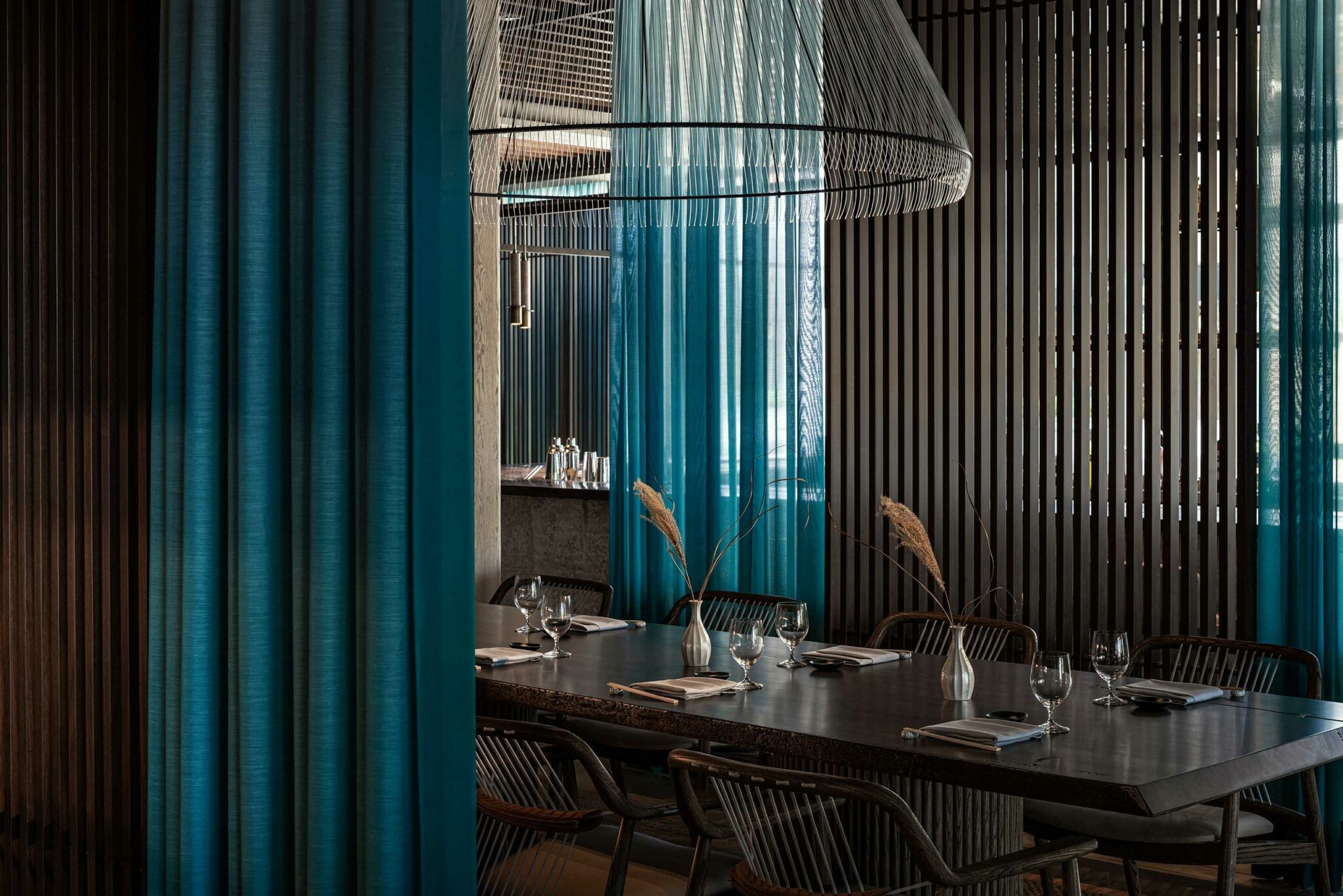 A private dining area at Tapasake, One&Only Portonovi, defined by teal drapery, vertical timber slats, sculptural wire lighting, and a dark timber table set with minimalist place settings—showcasing intimate, moody hospitality design with layered texture and material elegance.