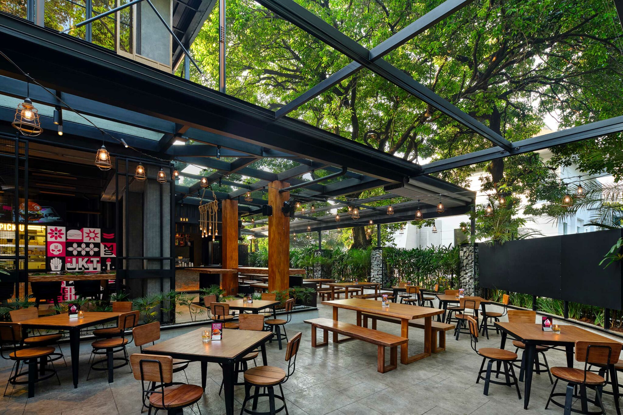 Open-air dining terrace at Meatsmith Jakarta featuring timber tables, industrial steel pergola, hanging lights, and tropical greenery