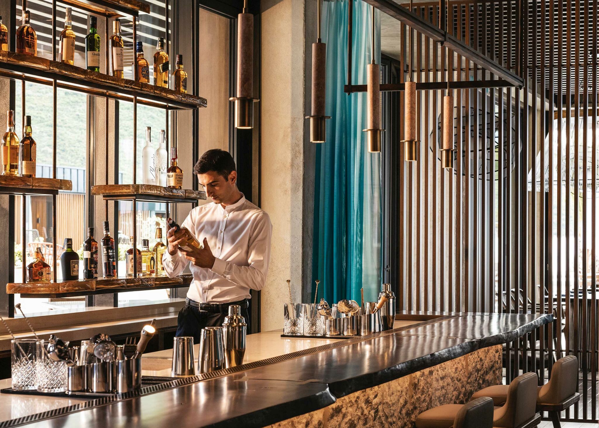 A bartender at Tapasake, One&Only Portonovi examines a bottle behind a sculpted stone bar, framed by vertical timber slats, teal glass accents, and warmly lit suspended shelving
