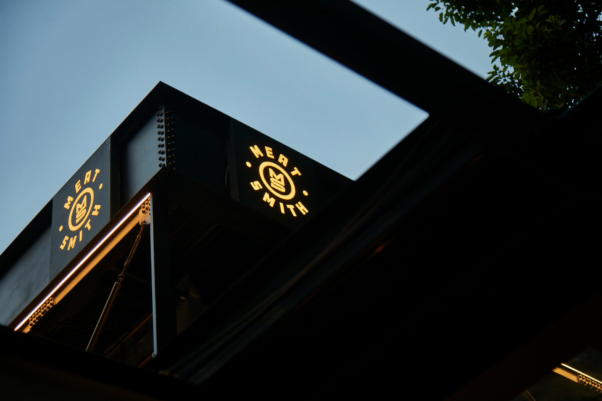 Illuminated Meatsmith logo sign glowing at dusk on a modern industrial façade.