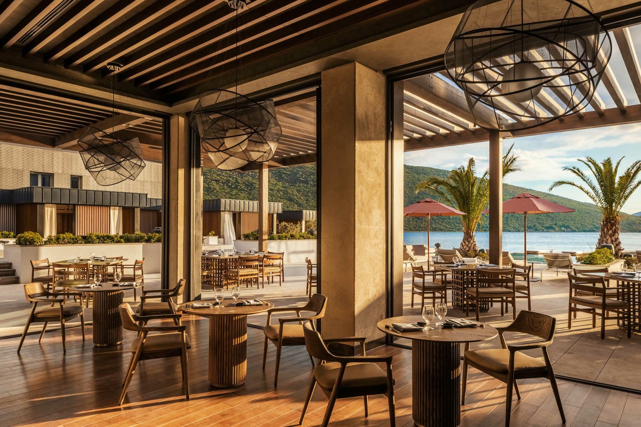Indoor–outdoor dining at Tapasake, One&Only Portonovi, featuring timber flooring, sculptural pendant lights, and open-air terraces overlooking the bay with palm trees and crimson umbrellas—showcasing elegant coastal hospitality design with a warm, sunset atmosphere.