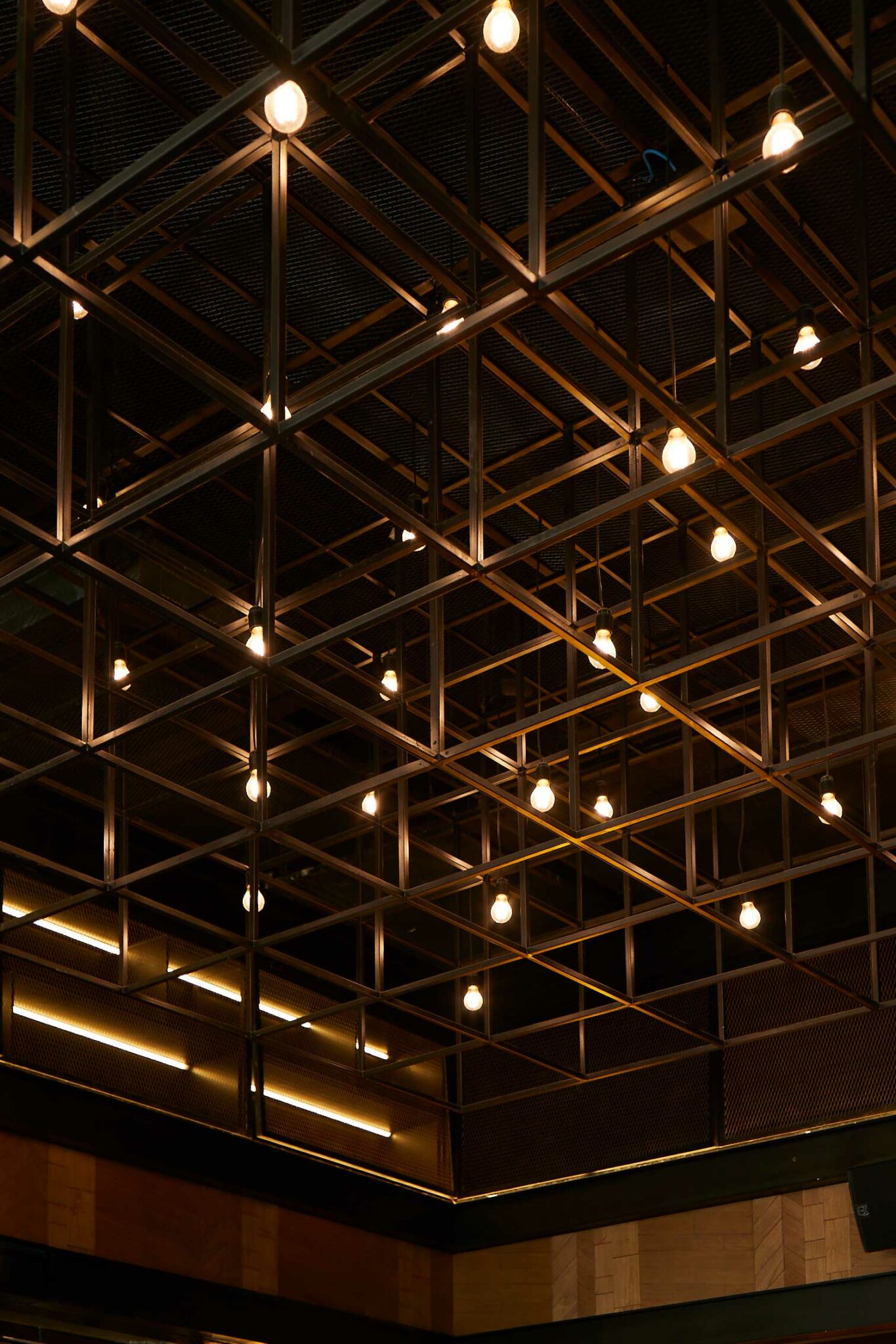 Geometric metal ceiling grid with exposed warm bulbs creating an industrial ambient glow.