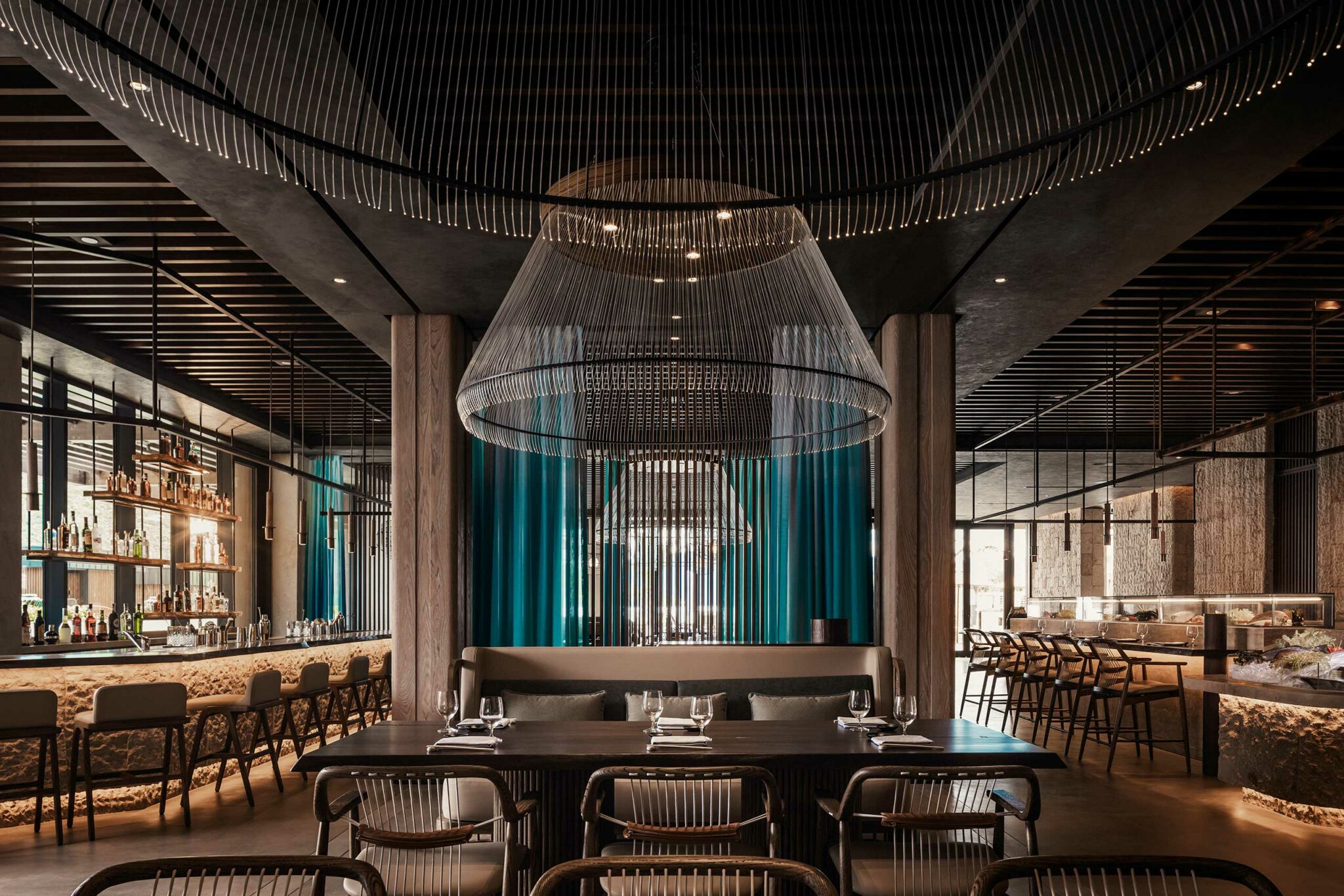 The interior of Tapasake at One&Only Portonovi, featuring sculptural wire chandeliers, a stone-clad bar, timber ceiling slats, and teal glass accents—showcasing refined, contemporary hospitality design with a strong sense of materiality, rhythm, and coastal elegance.