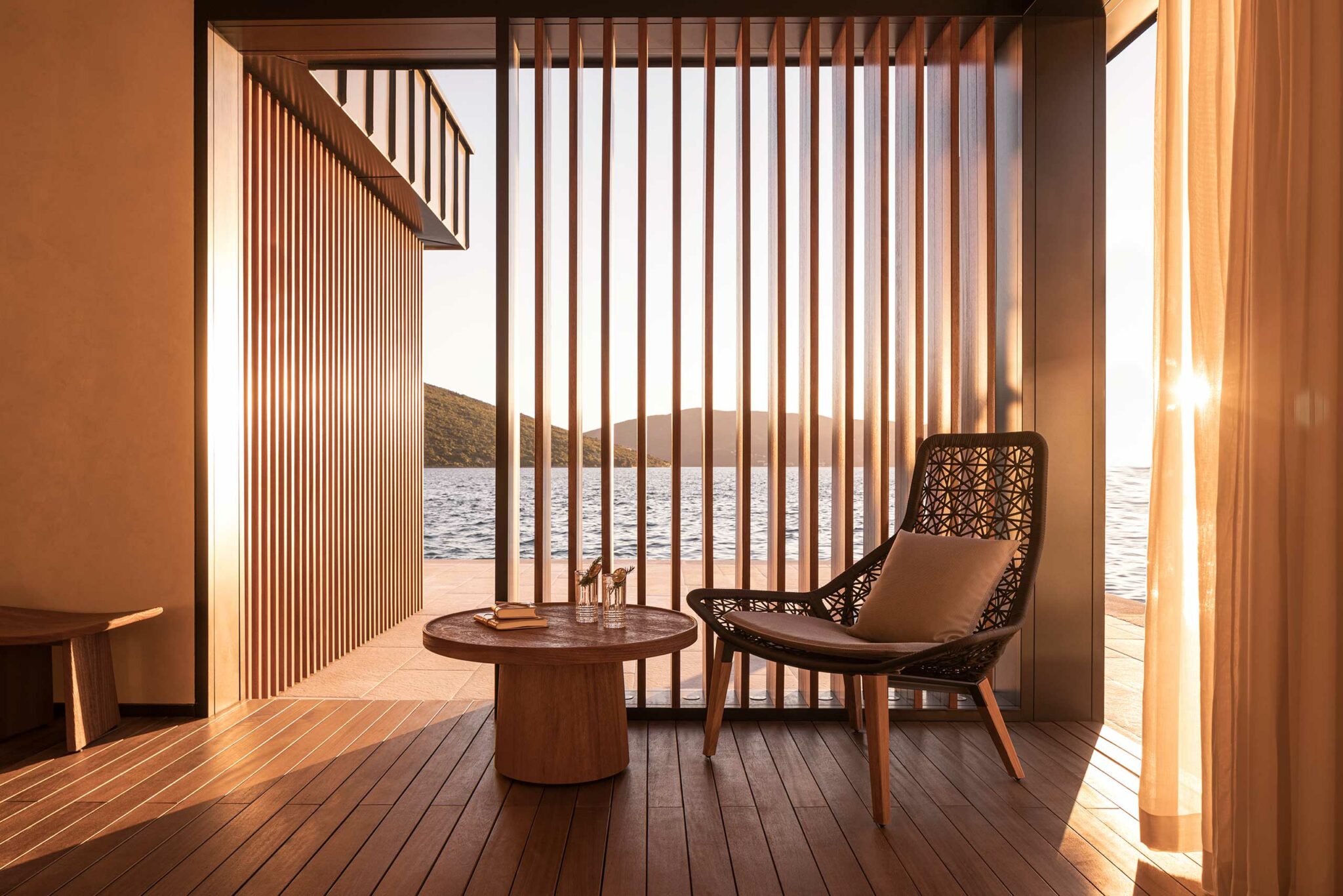 A serene terrace at Tapasake, One&Only Portonovi, featuring warm timber decking, vertical slatted screens, and a woven lounge chair overlooking the bay at sunset—highlighting refined, indoor–outdoor hospitality design with a calm, coastal sensibility.