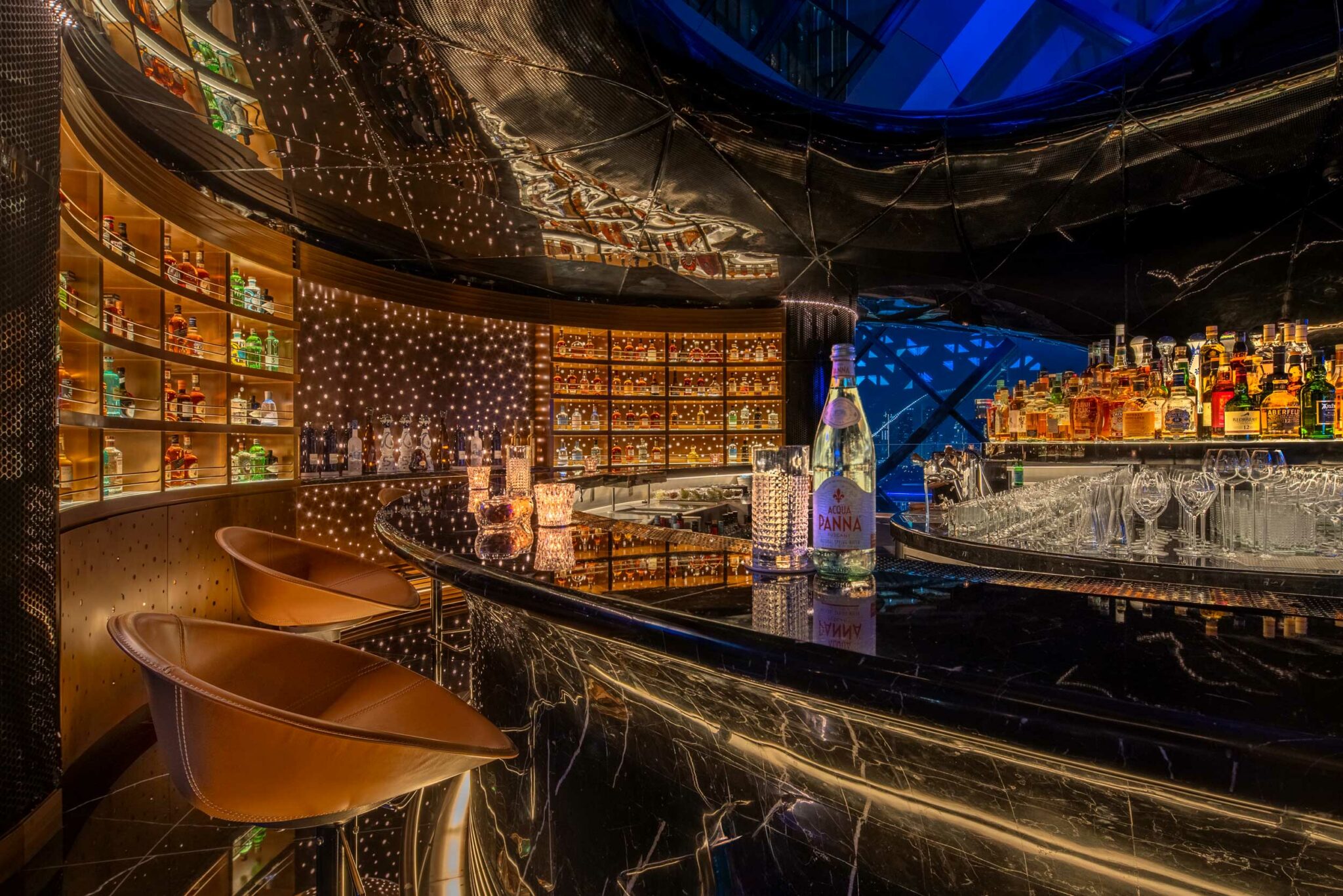 Interior of Sphere at One Za’abeel Dubai, featuring a glossy black marble bar surrounded by warm backlit bottle displays, star-like pin lights across the curved wall, and panoramic night views reflected in the metallic ceiling.