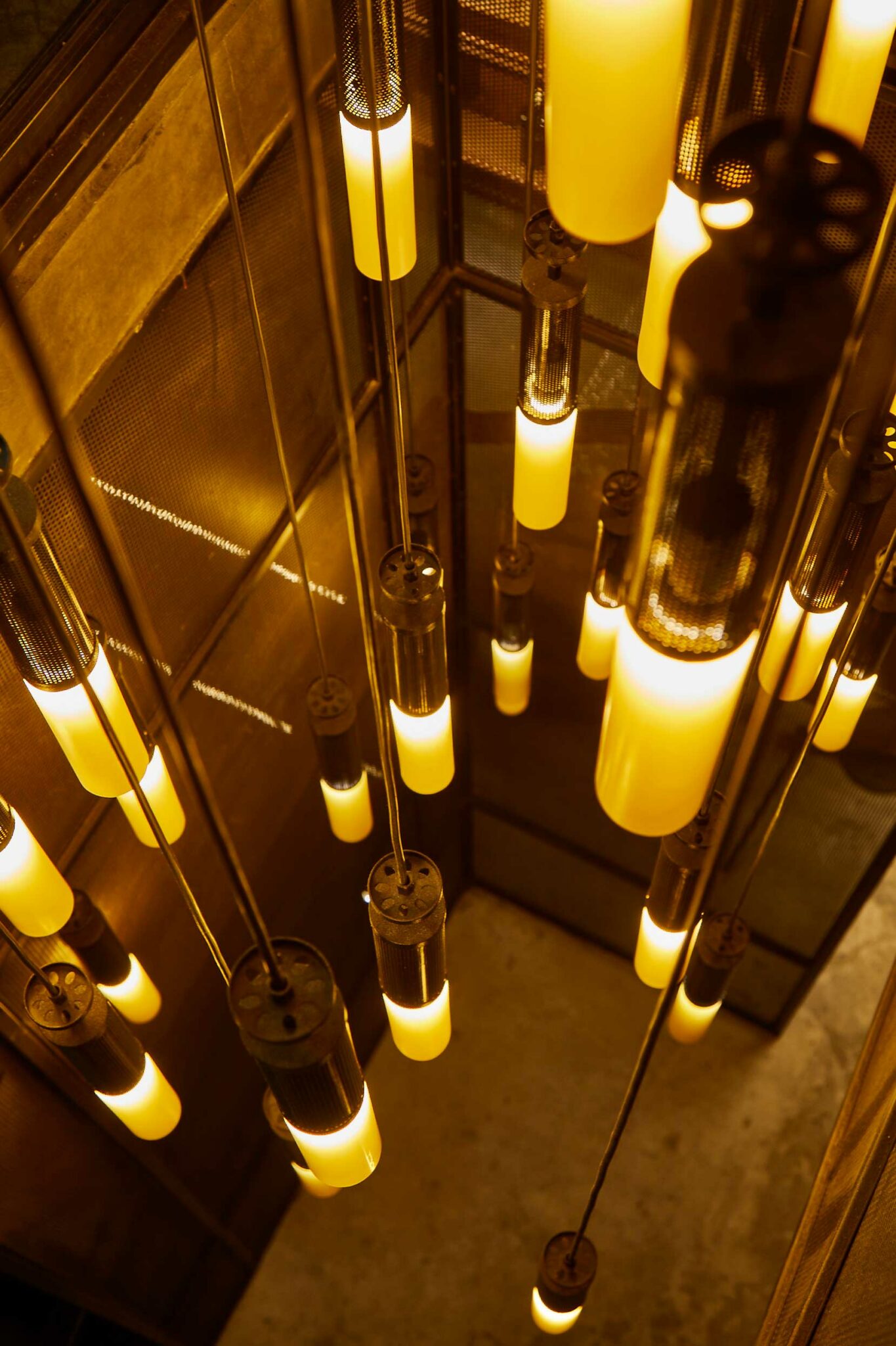 A cluster of cylindrical pendant lights suspended at varying heights, glowing in warm amber tones against perforated metal panels