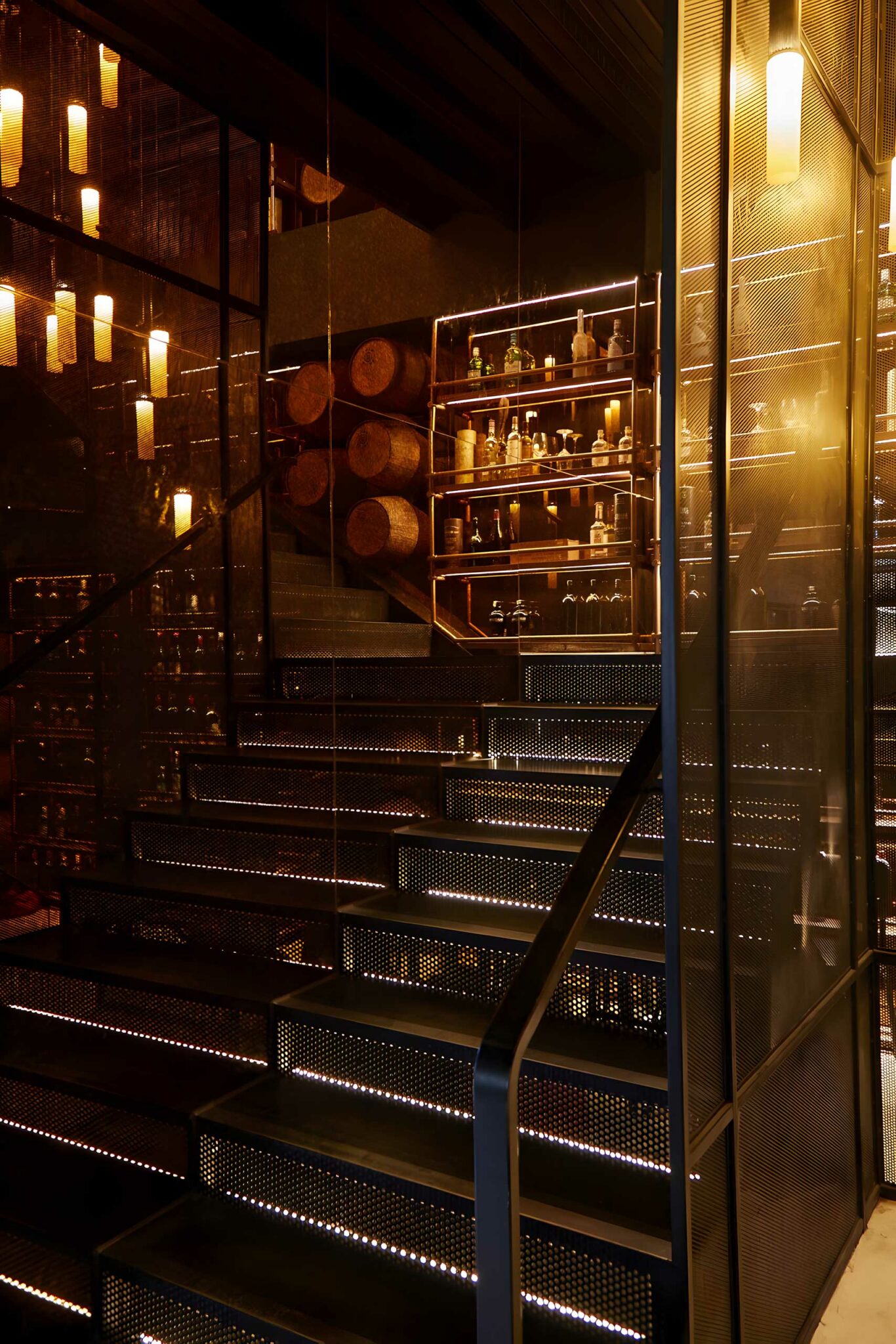 A dramatic staircase at Meat Smith Jakarta, illuminated with embedded step lighting and framed by perforated metal, with shelves of spirits and stacked wooden barrels visible through warm, reflective glass—showcasing an industrial, atmospheric hospitality design rooted in craft and detail.