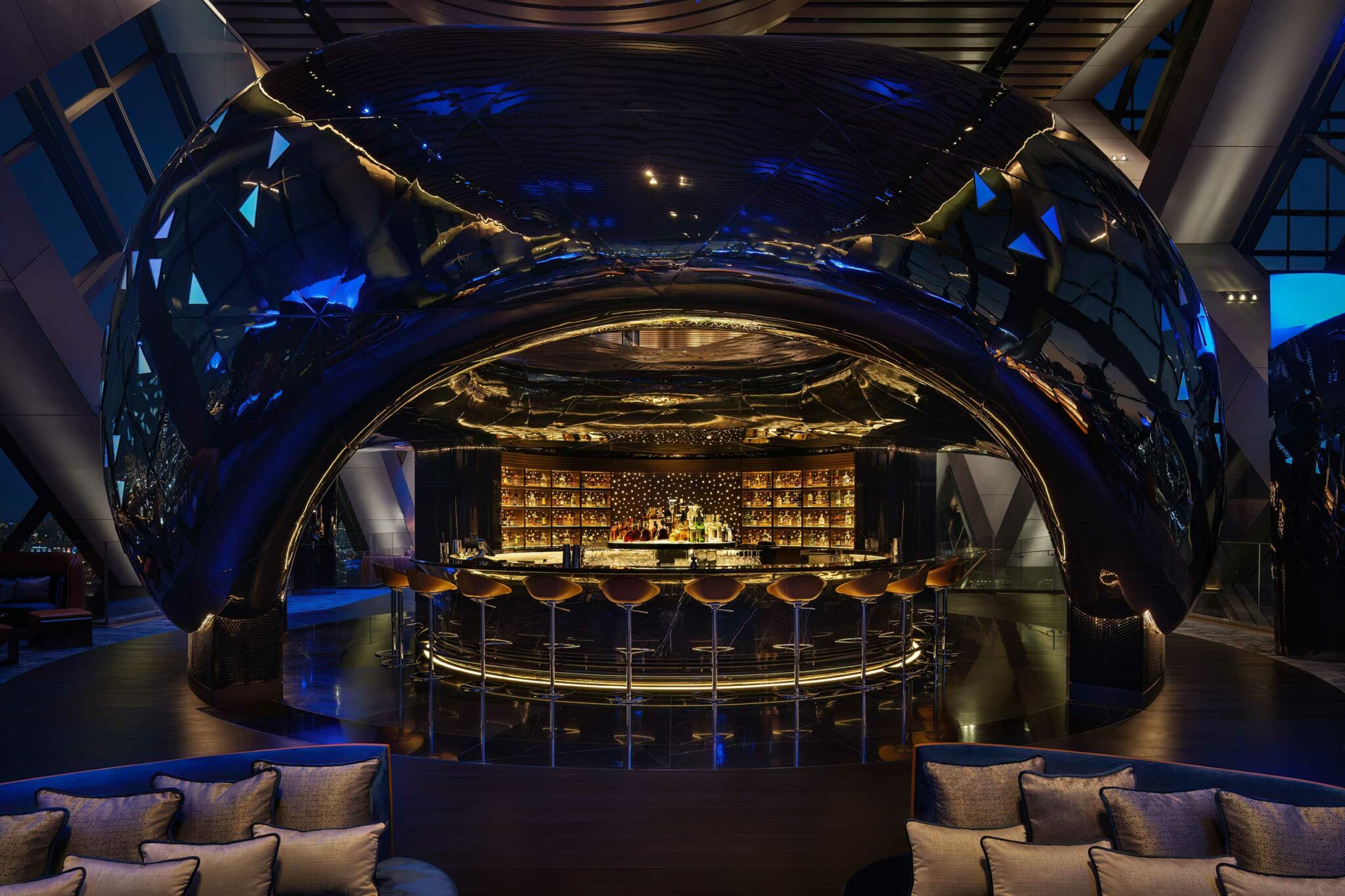 Dramatic frontal view of the circular bar enveloped by the cavernous, mirror-black sculptural form of Sphere; warm golden backlighting highlights the bottle display while the blue exterior lighting reflects across the metallic surfaces.