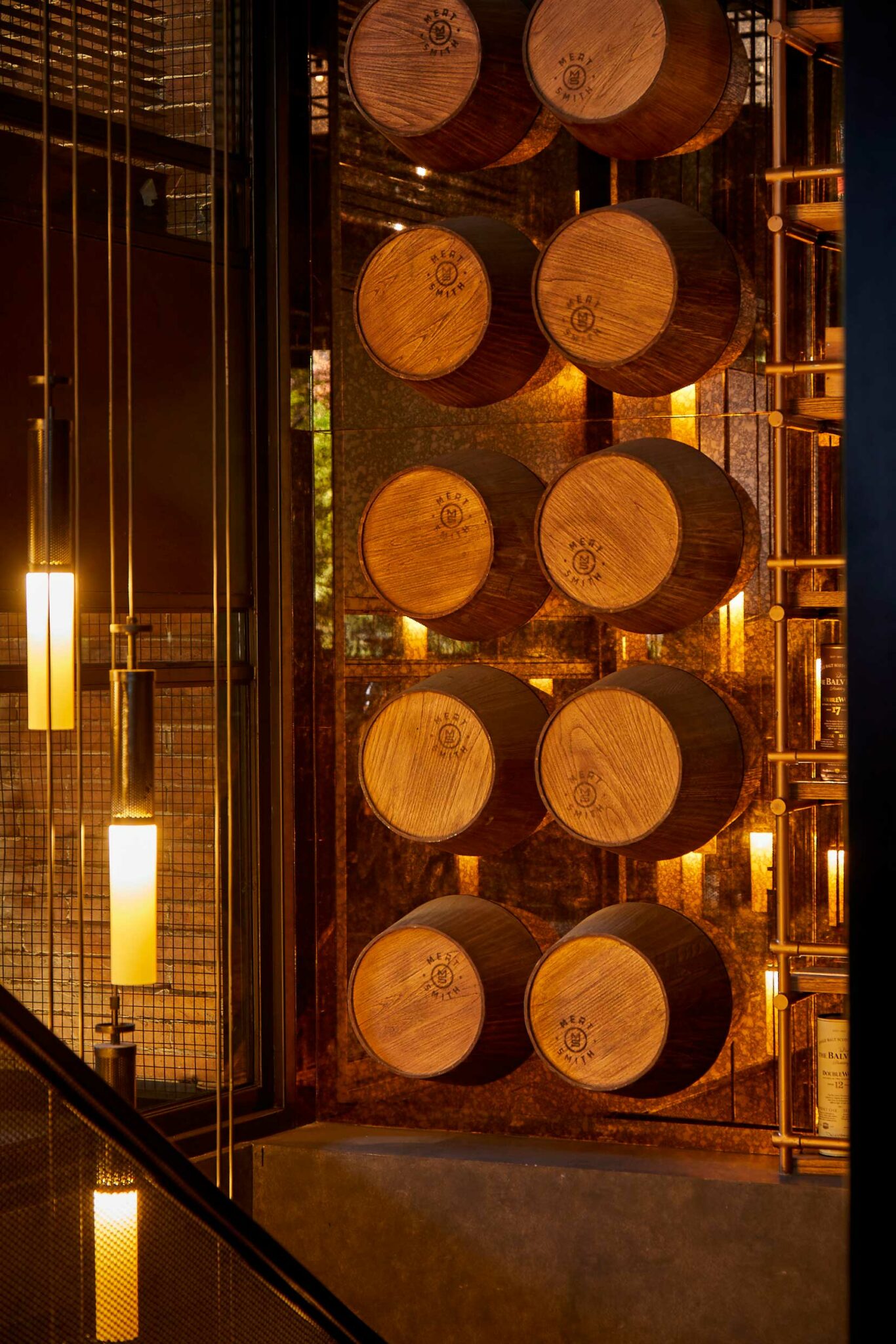 A feature wall at Meat Smith Jakarta displaying stacked wooden barrels stamped with the restaurant’s logo, illuminated by warm pendant lighting and framed by industrial metalwork—showcasing a bold, craft-centred hospitality design with strong material character.