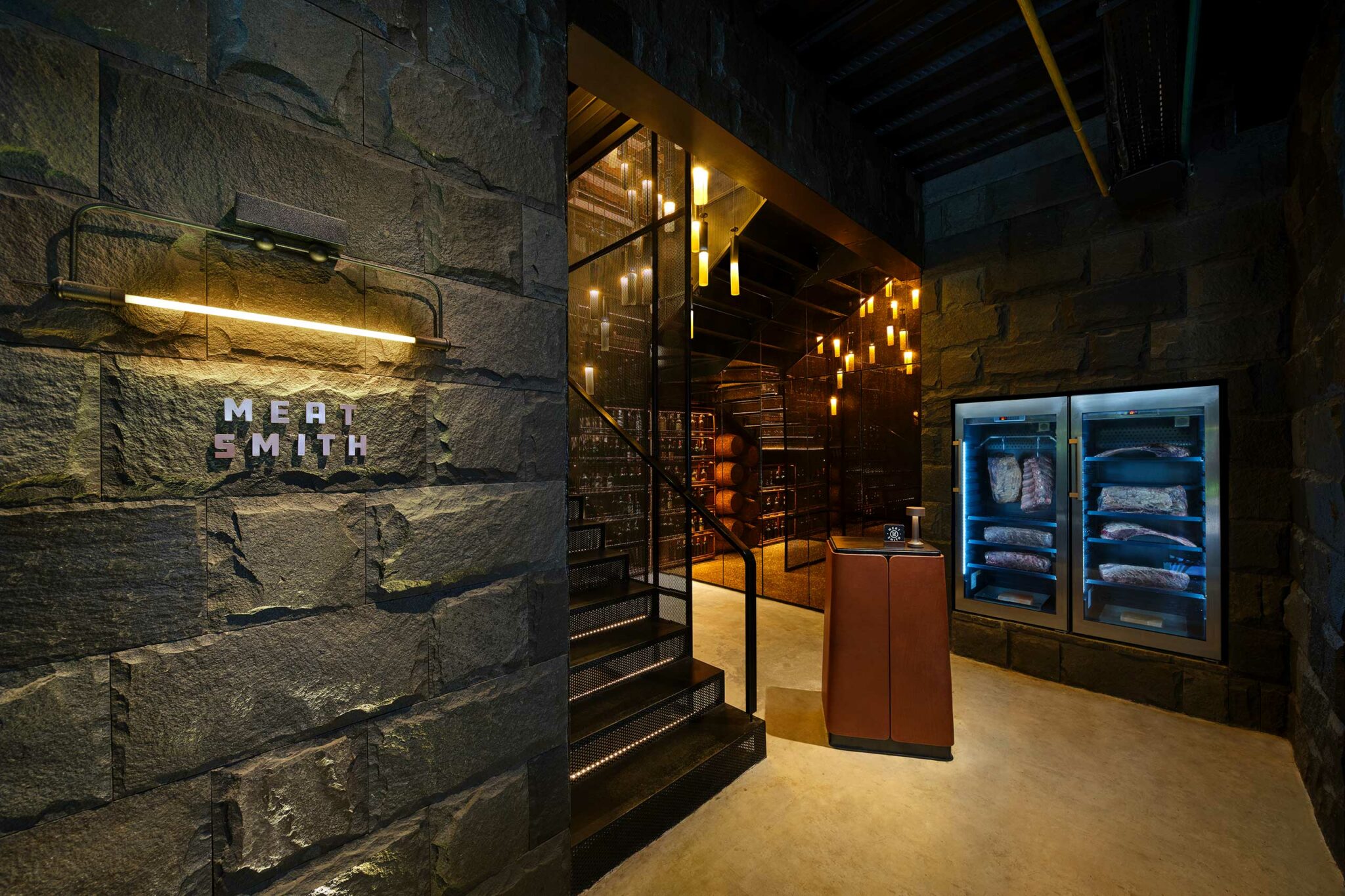 The entrance to Meat Smith, featuring rugged stone walls, illuminated signage, a glass-walled staircase leading to a warmly lit cellar, and refrigerated dry-age cabinets displaying cuts of meat.