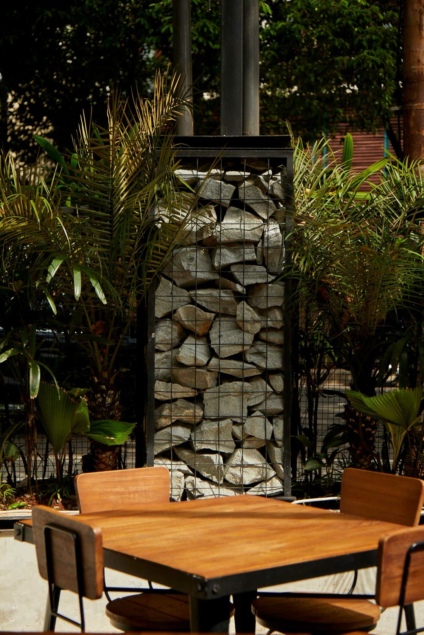 An outdoor dining area with a simple wooden table and chairs set beside a gabion-style wall filled with stacked stones, surrounded by lush tropical plants—showcasing a raw, material-driven approach to casual hospitality design that blends nature and industrial elements.