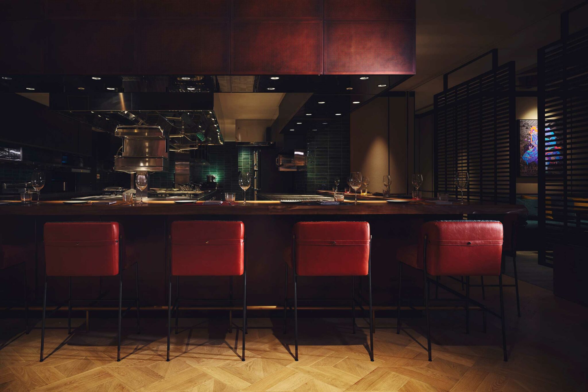 A moody chef’s counter at Gallery 11 with deep red leather bar stools, a polished wooden countertop, and an open kitchen framed by dark metal and green-glazed tiles.