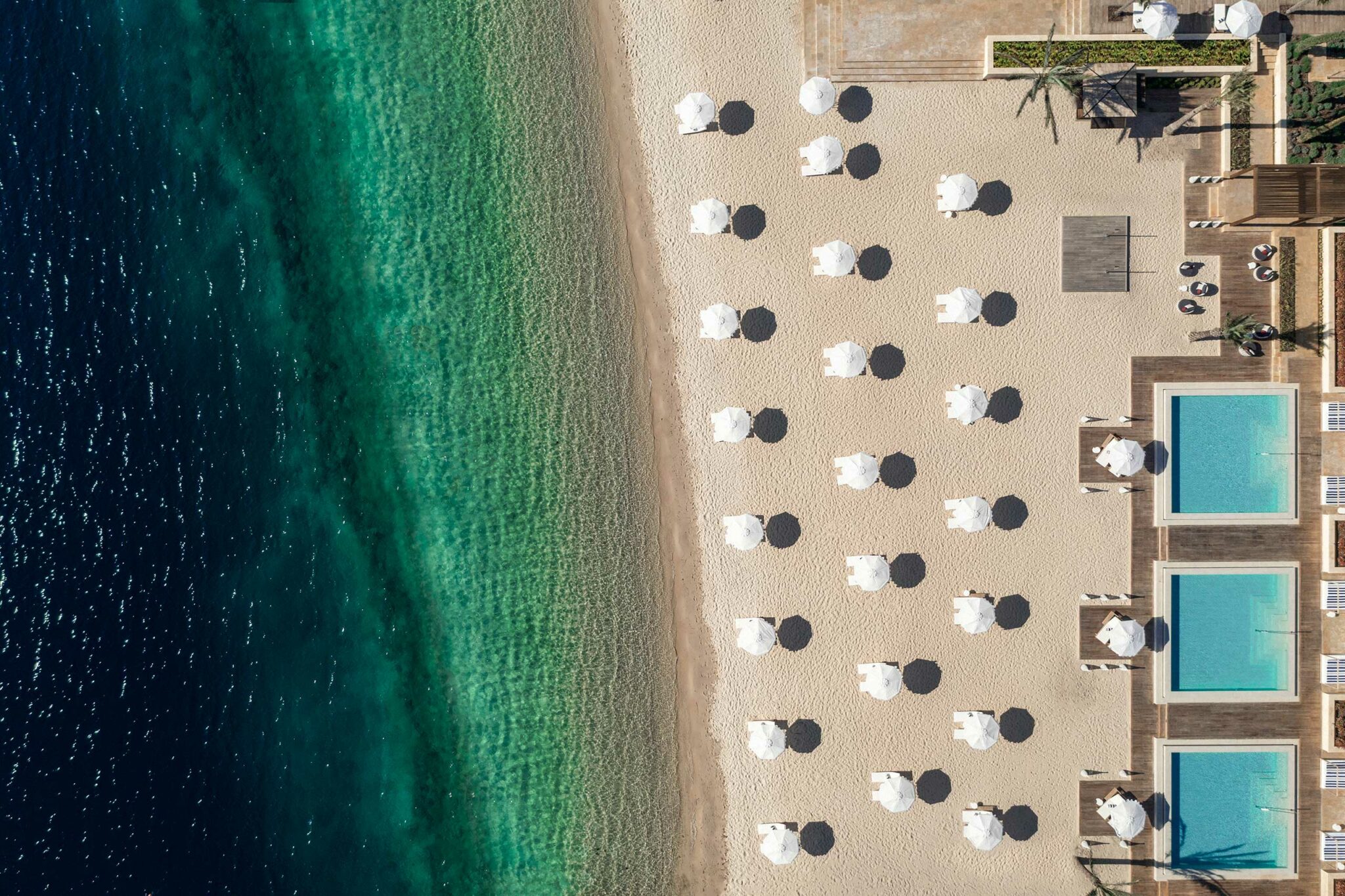 Top-down view of a pristine beach with rows of white umbrellas casting circular shadows, clear turquoise water on one side, and a sequence of geometric pools on the other—showcasing meticulous resort planning and coastal hospitality design.
