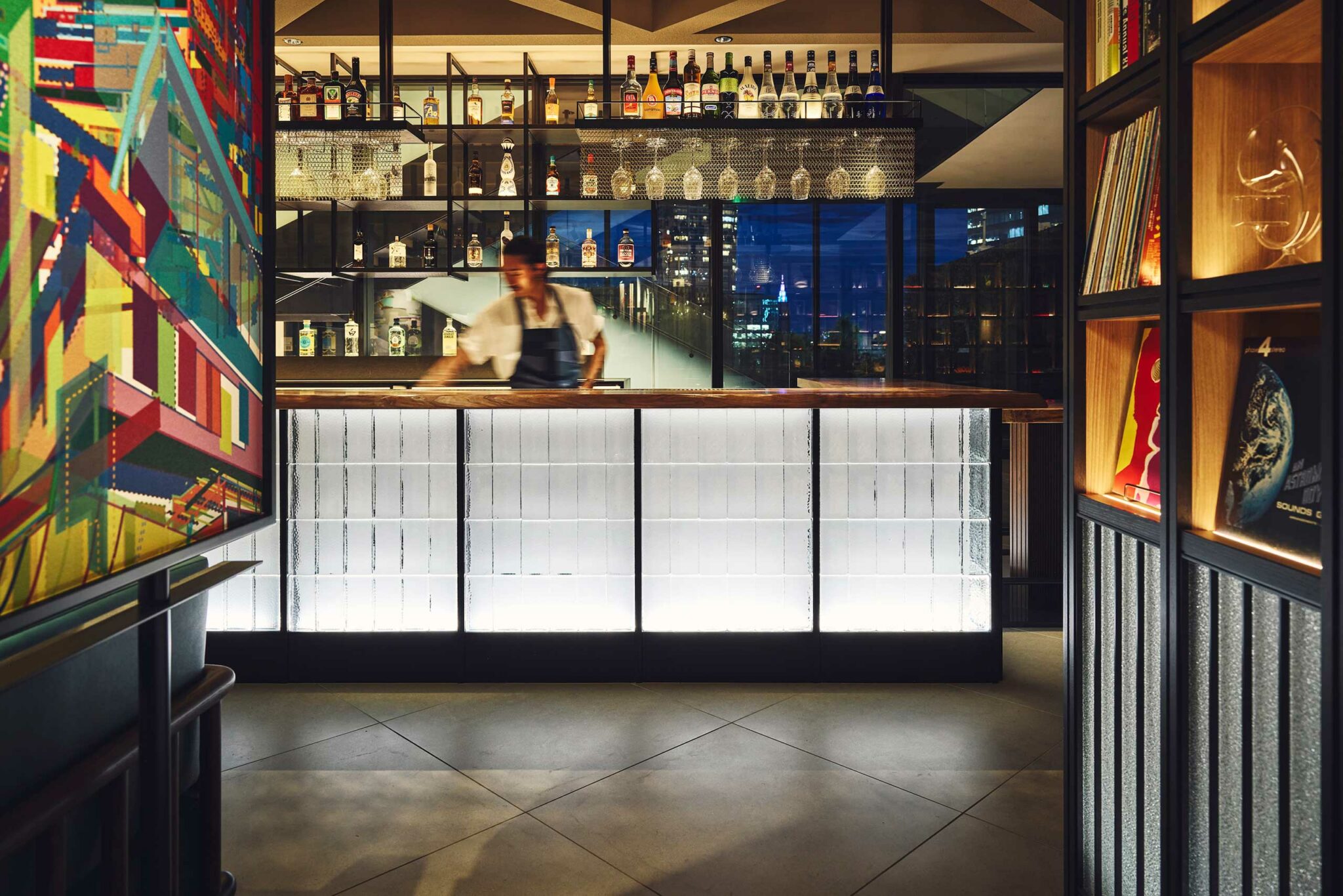 A contemporary bar at Gallery 11 in Hotel Indigo Shibuya, featuring a backlit glass-block counter, suspended shelves displaying premium spirits, and a blurred bartender in motion against a backdrop of city lights.