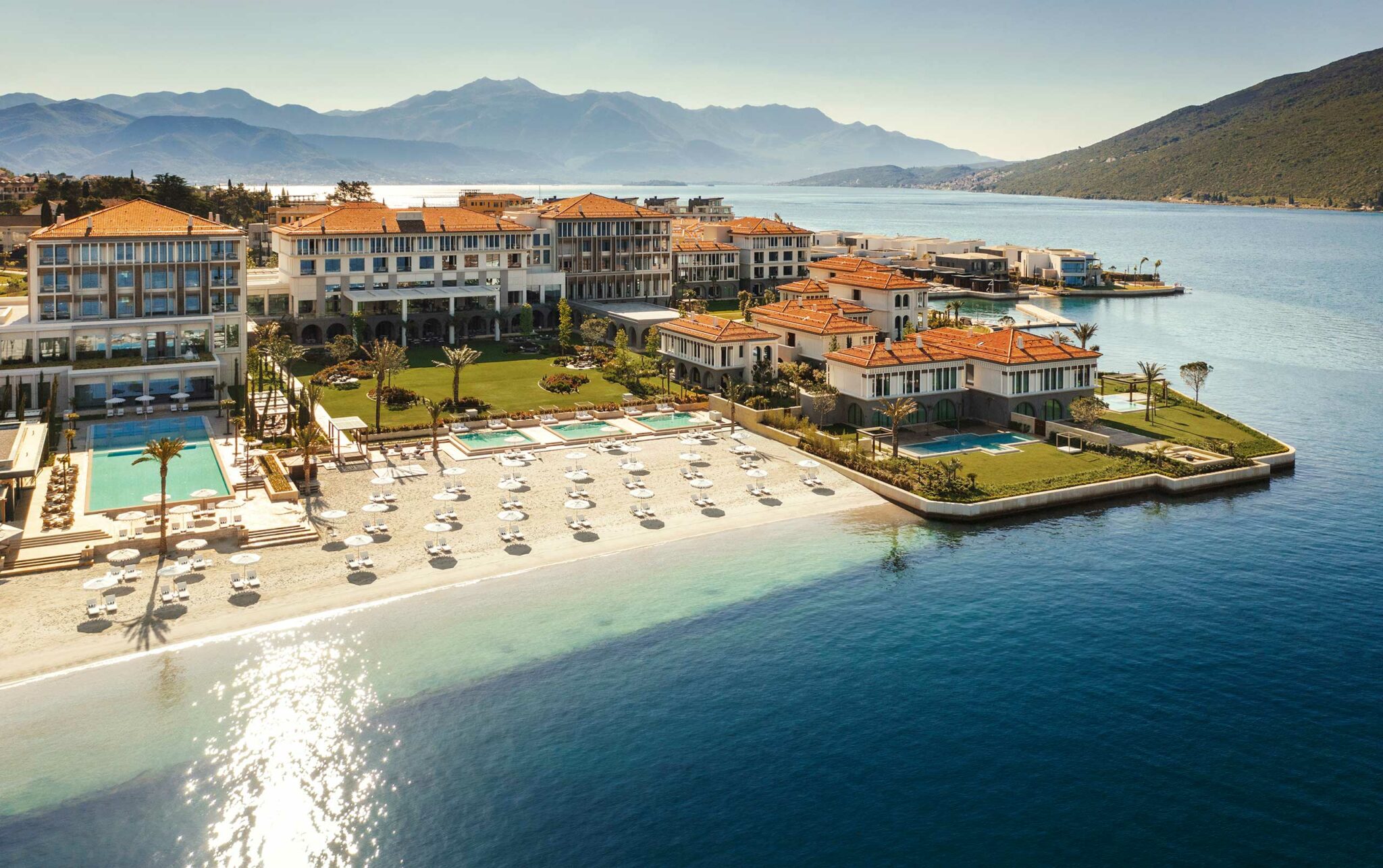Aerial view of a luxury waterfront resort with terracotta-roofed buildings, pools, landscaped gardens, and a private beach lined with umbrellas, set against a calm bay and mountain backdrop—illustrating large-scale, resort-level hospitality design in a scenic coastal setting.