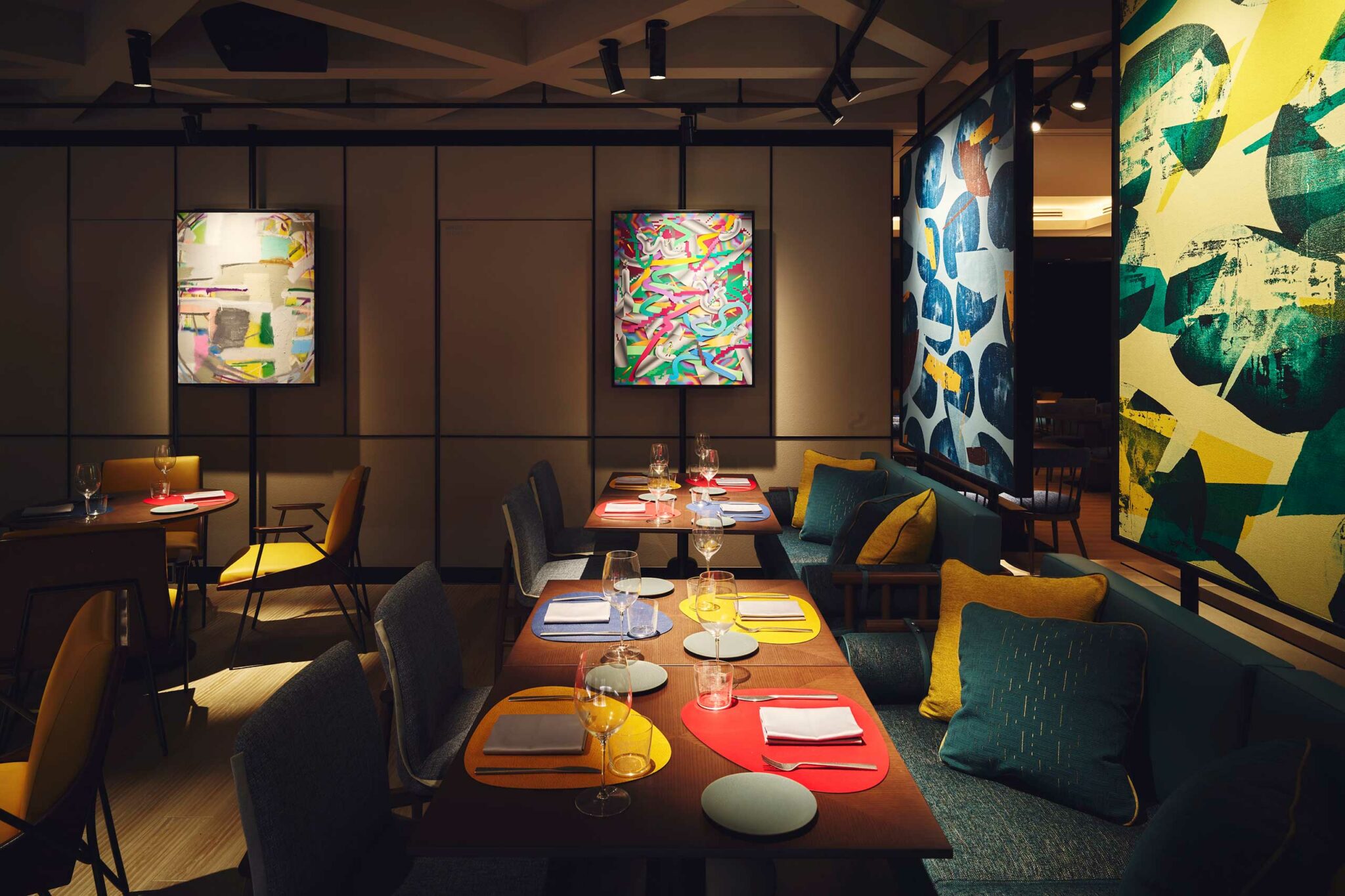 A colourful dining room featuring abstract art panels and framed contemporary pieces, with tables set with vibrant placemats in red, yellow, and blue, alongside cosy upholstered seating and decorative cushions.