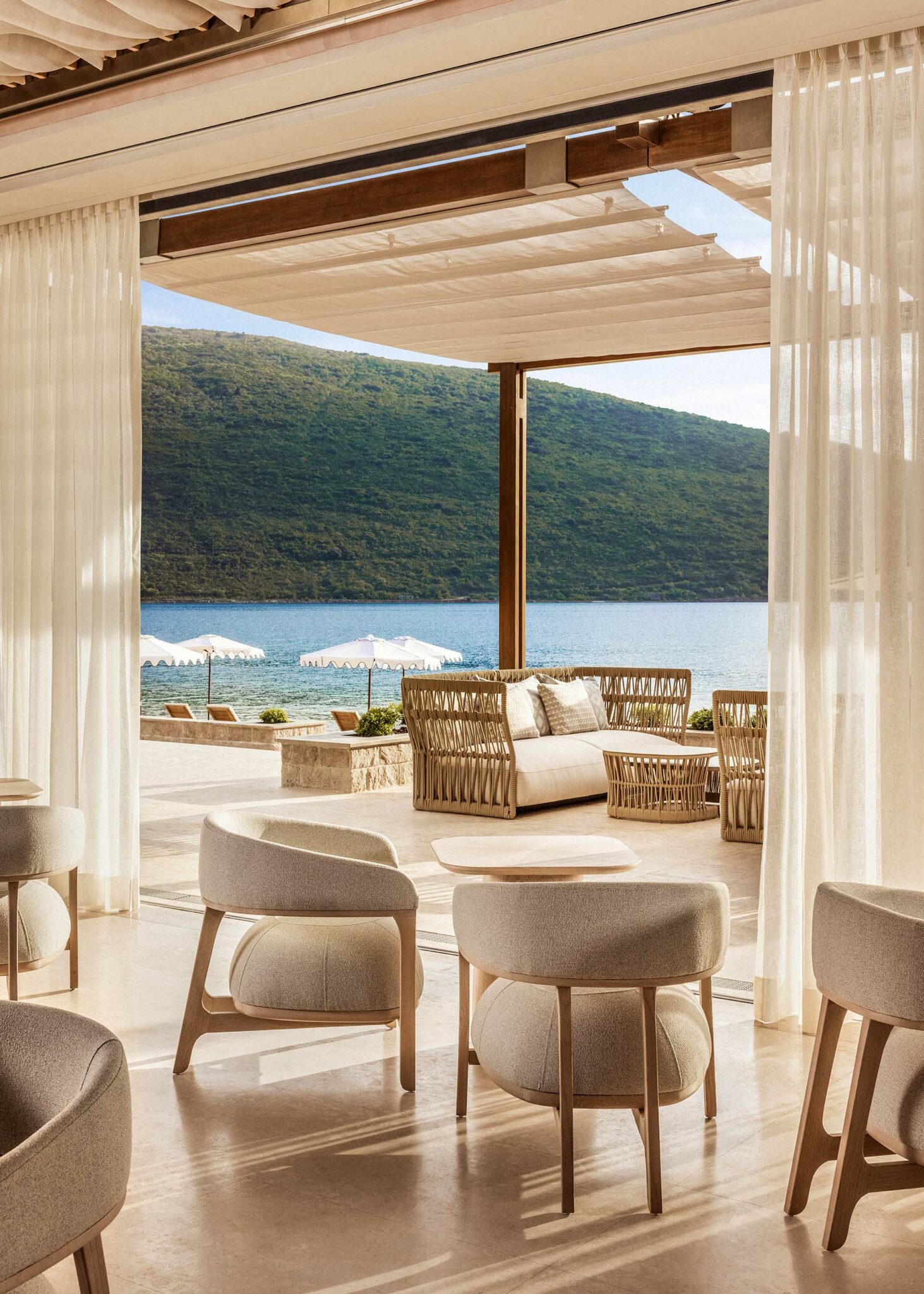 An airy indoor–outdoor lounge with soft neutral furnishings, sheer curtains, and woven terrace seating overlooking a calm bay and green hillside—expressing a serene, resort-style hospitality environment designed for relaxed coastal dining.