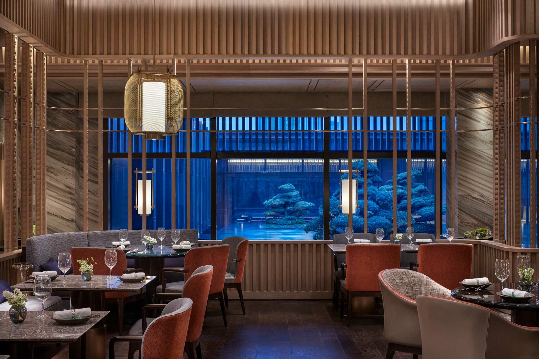 Elegant dining room at Park Hyatt Suzhou featuring marble tables, red and grey upholstered chairs, decorative wooden screens, and large windows overlooking a serene courtyard with sculpted bonsai trees. Inspired by the grand halls and pavilions of suzhou