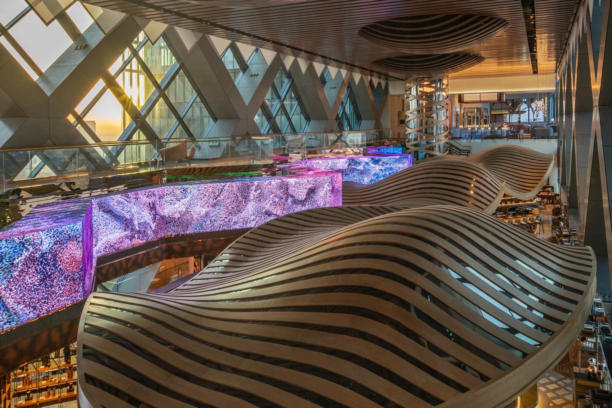 Dramatic overhead view of Arrazuna’s layered timber wave structures and expansive LED screens displaying colourful abstract textures beneath the glass façade