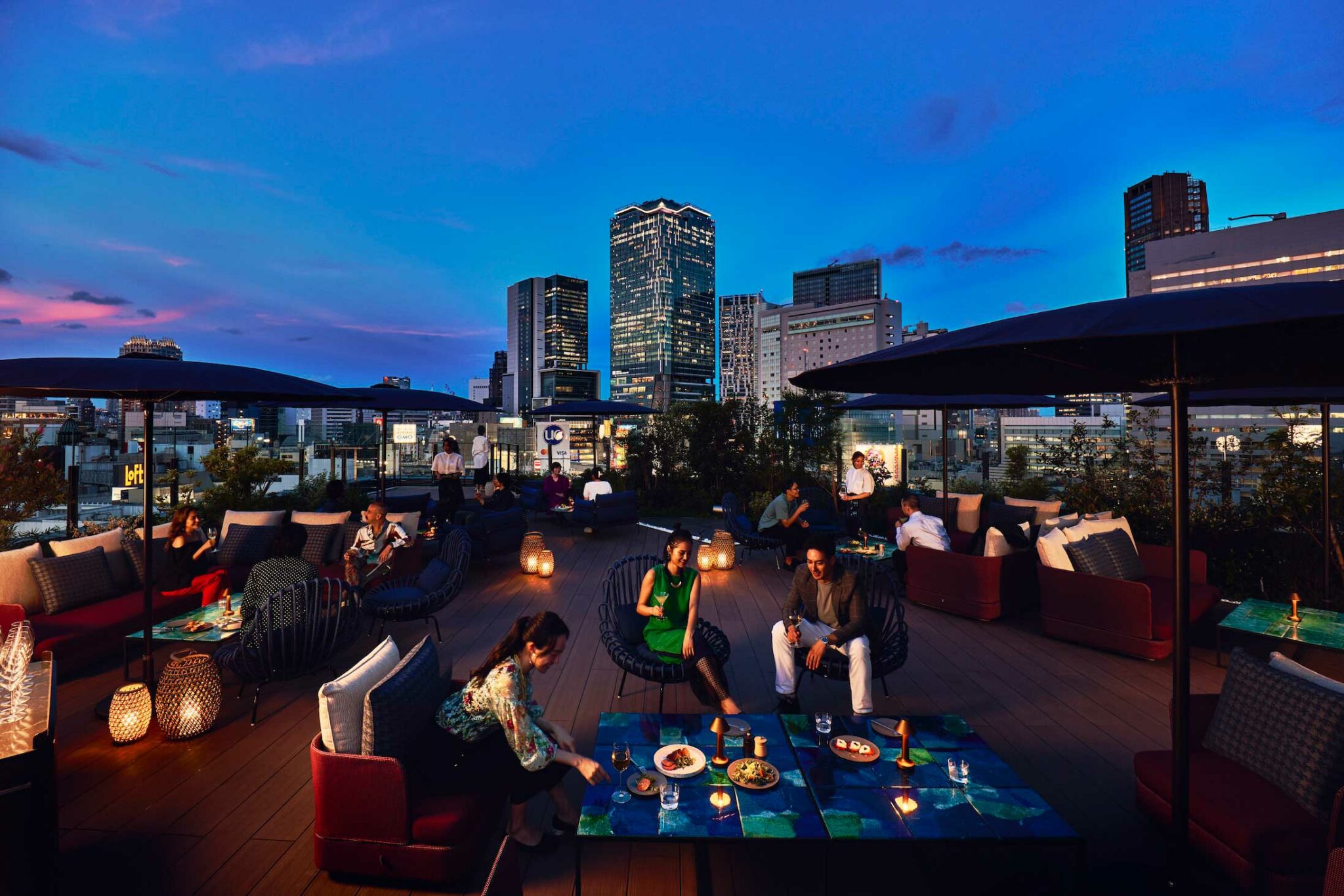 A rooftop terrace at dusk overlooking Tokyo’s skyline, with guests relaxing on modern outdoor sofas under umbrellas, sharing small plates and drinks in a lively, atmospheric setting.
