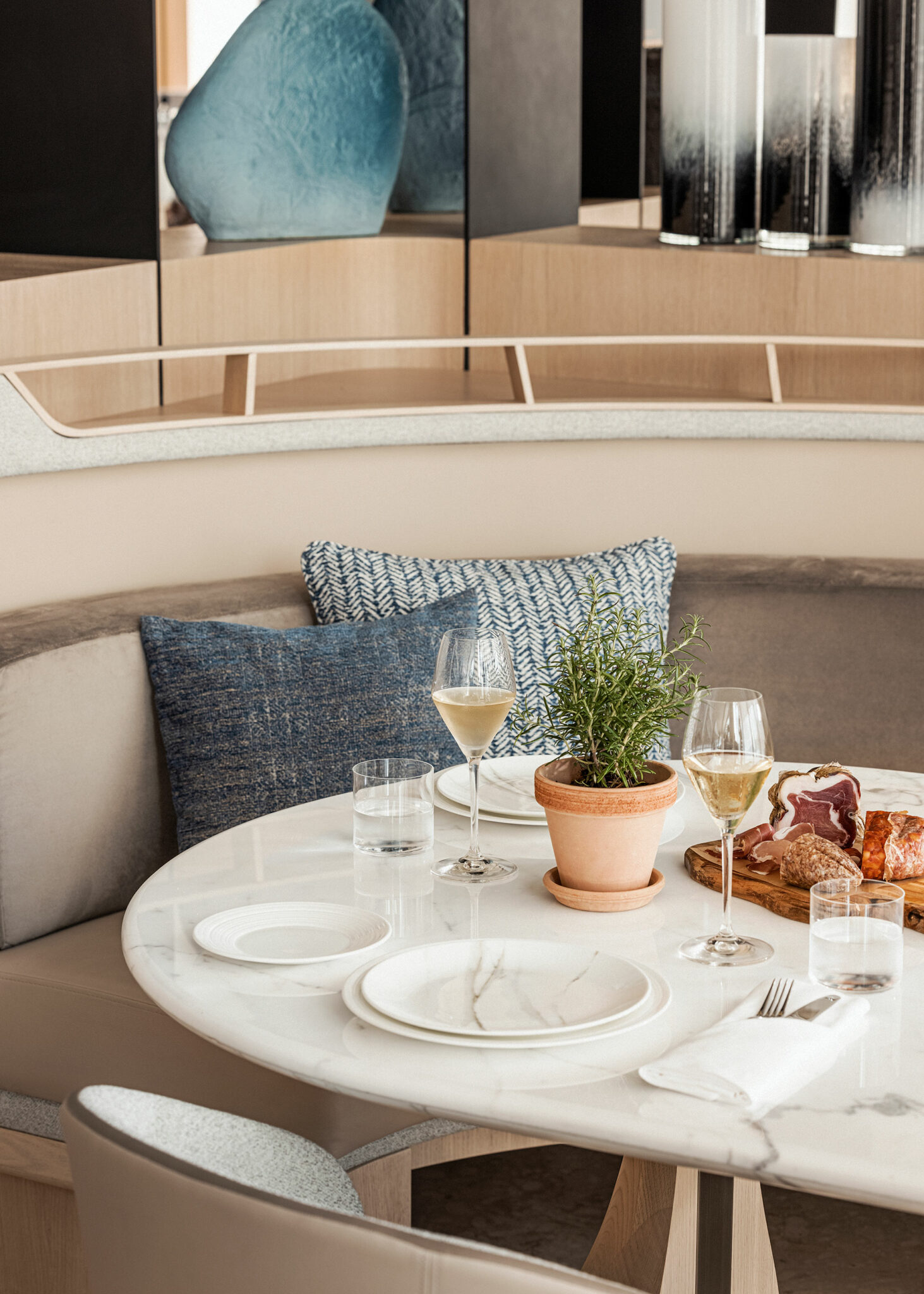 A marble-topped round dining table set with white plates, wine glasses, and a terracotta pot of rosemary, beside a curved banquette with textured blue cushions and sculptural décor—highlighting a refined, contemporary hospitality interior crafted for relaxed, elevated dining.