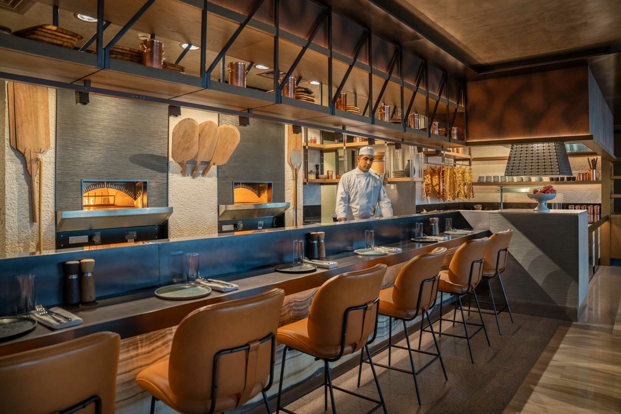 Open kitchen counter at Arrazuna showcasing wood-fired ovens, hanging wooden peels, copper cookware, and a chef working behind a row of tan leather counter stools.