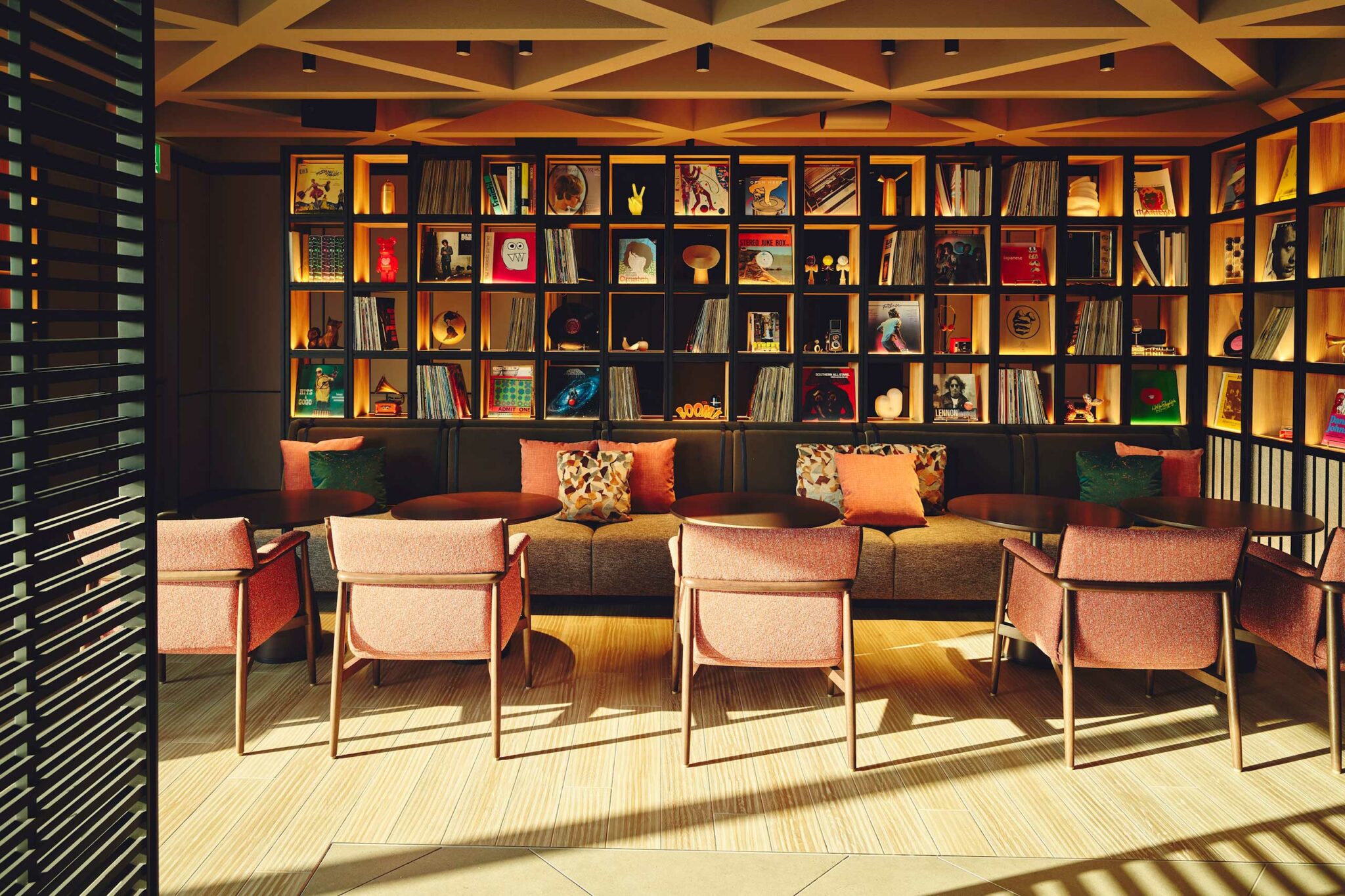 A lounge-style seating area with mid-century modern chairs and sofas facing a full wall of illuminated shelves filled with vinyl records, books, and eclectic art objects, bathed in soft golden afternoon light.
