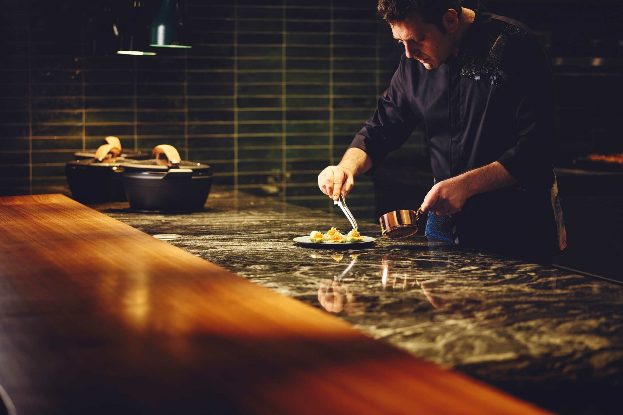 A chef in Gallery 11 Hotel Indigo Tokyo Shibuya in a dark uniform meticulously plates a dish on a polished black-marble counter, with warm directional lighting highlighting the workspace and tiled dark-green backsplash behind him.