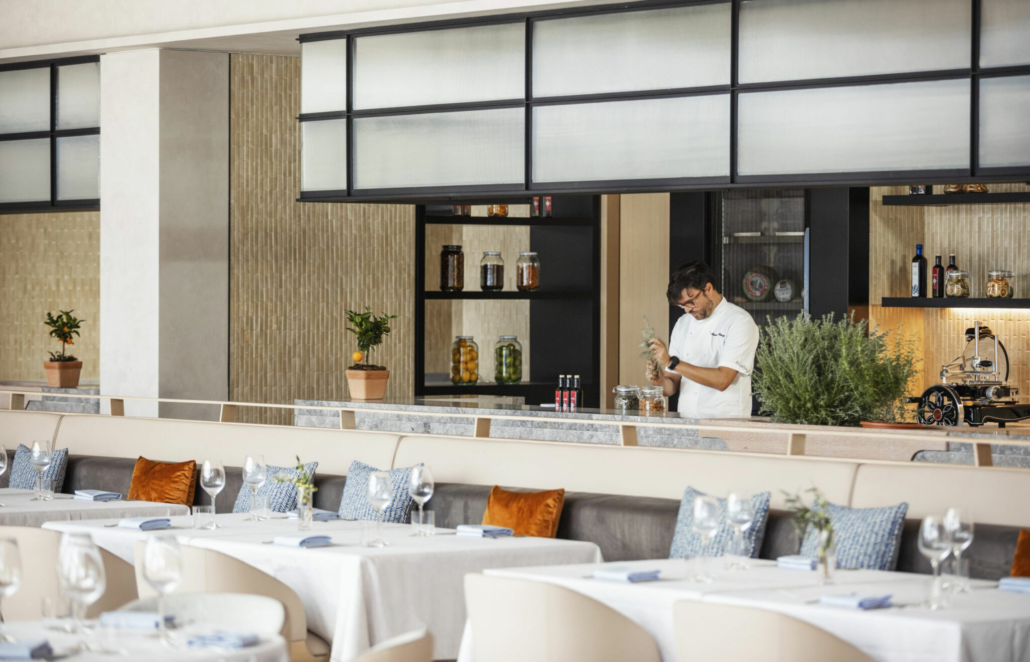 A light-filled restaurant interior featuring tailored seating, soft blue accents, and an open preparation counter where a chef works with herbs, capturing a crafted, detail-driven approach to modern hospitality design.
