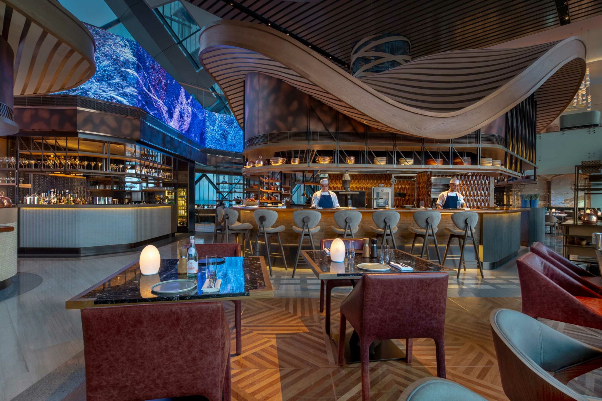 Arrazuna’s main dining space with sculptural wave-like ceiling forms, illuminated chef’s counter seating, and large digital screens displaying blue abstract visuals.
