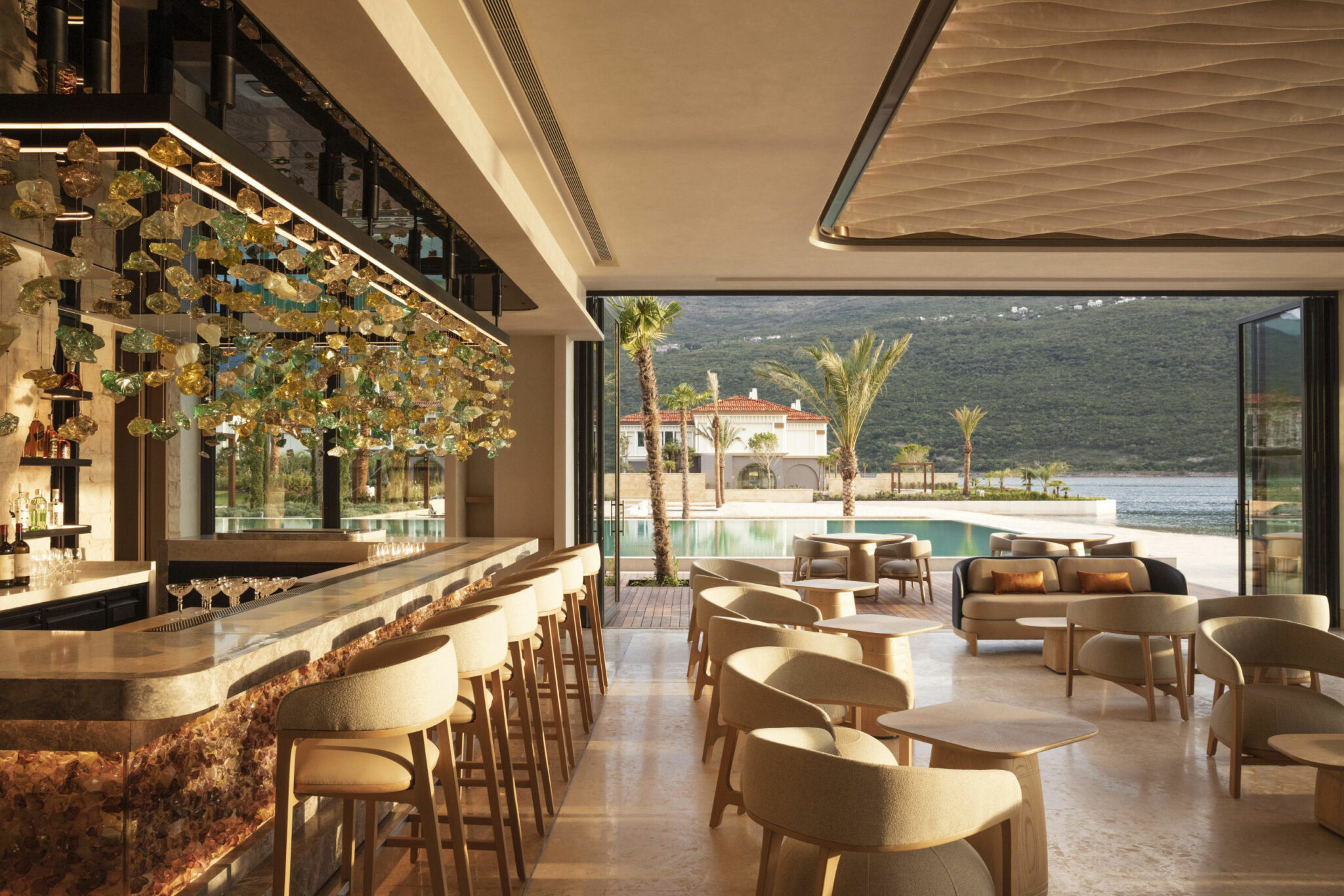 A luxurious open-air bar and lounge with soft beige seating, a long marble counter, and a sculptural ceiling installation of hanging glass ornaments, overlooking a pool, palm trees, and a coastal mountain landscape at sunset.