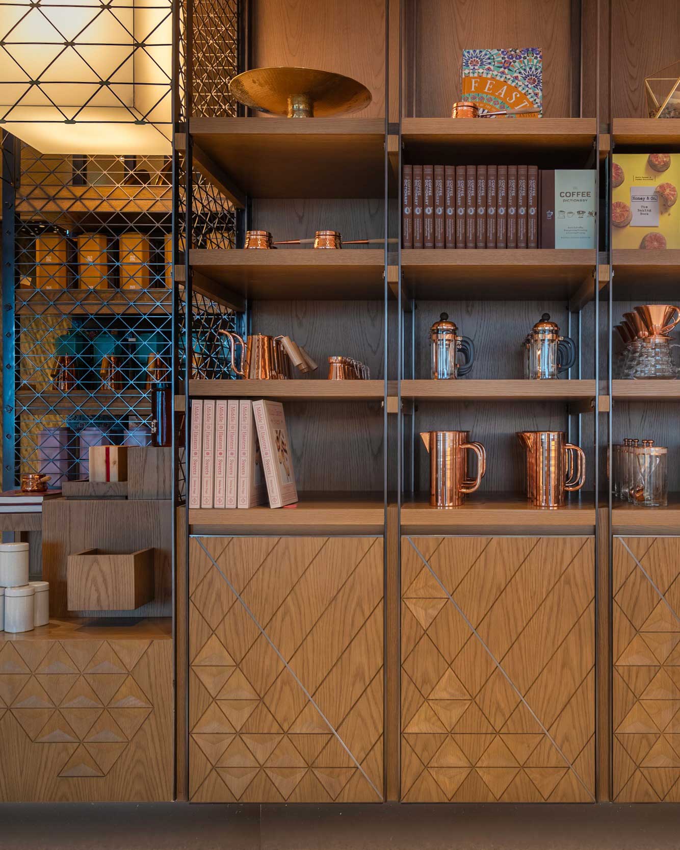 Stylish retail shelves at Arrazuna displaying copper coffee pots, glass jars, cookbooks, and geometric wood cabinetry, with soft lighting highlighting the warm, crafted materials.