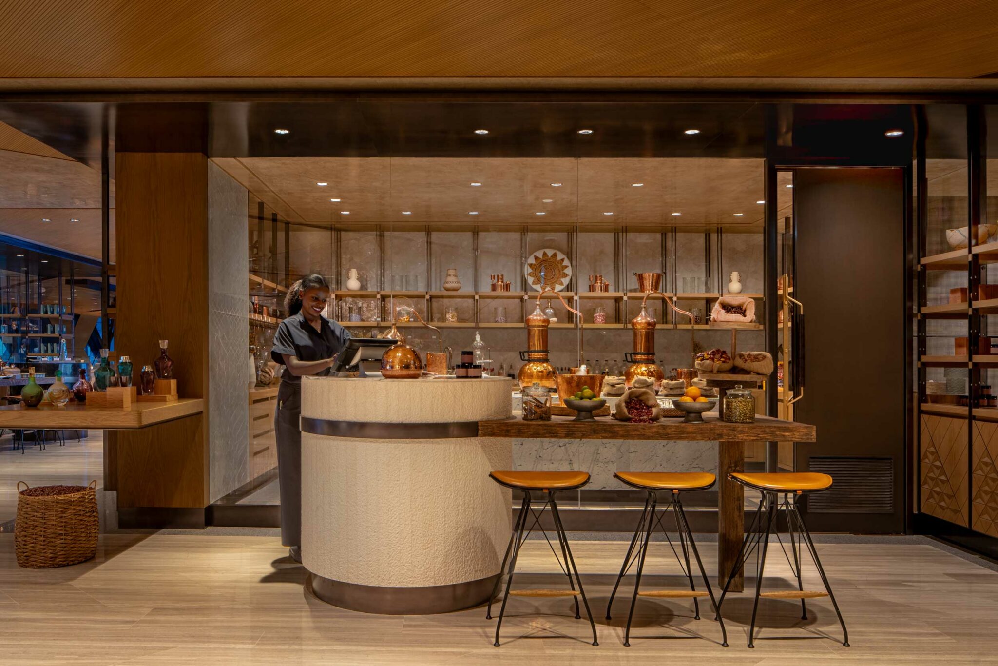 A staff member stands behind a rounded textured counter at Arrazuna, surrounded by copper distillation vessels, bowls of fresh produce, and warm wooden shelving displaying jars, ceramics, coffee,and ingredients.