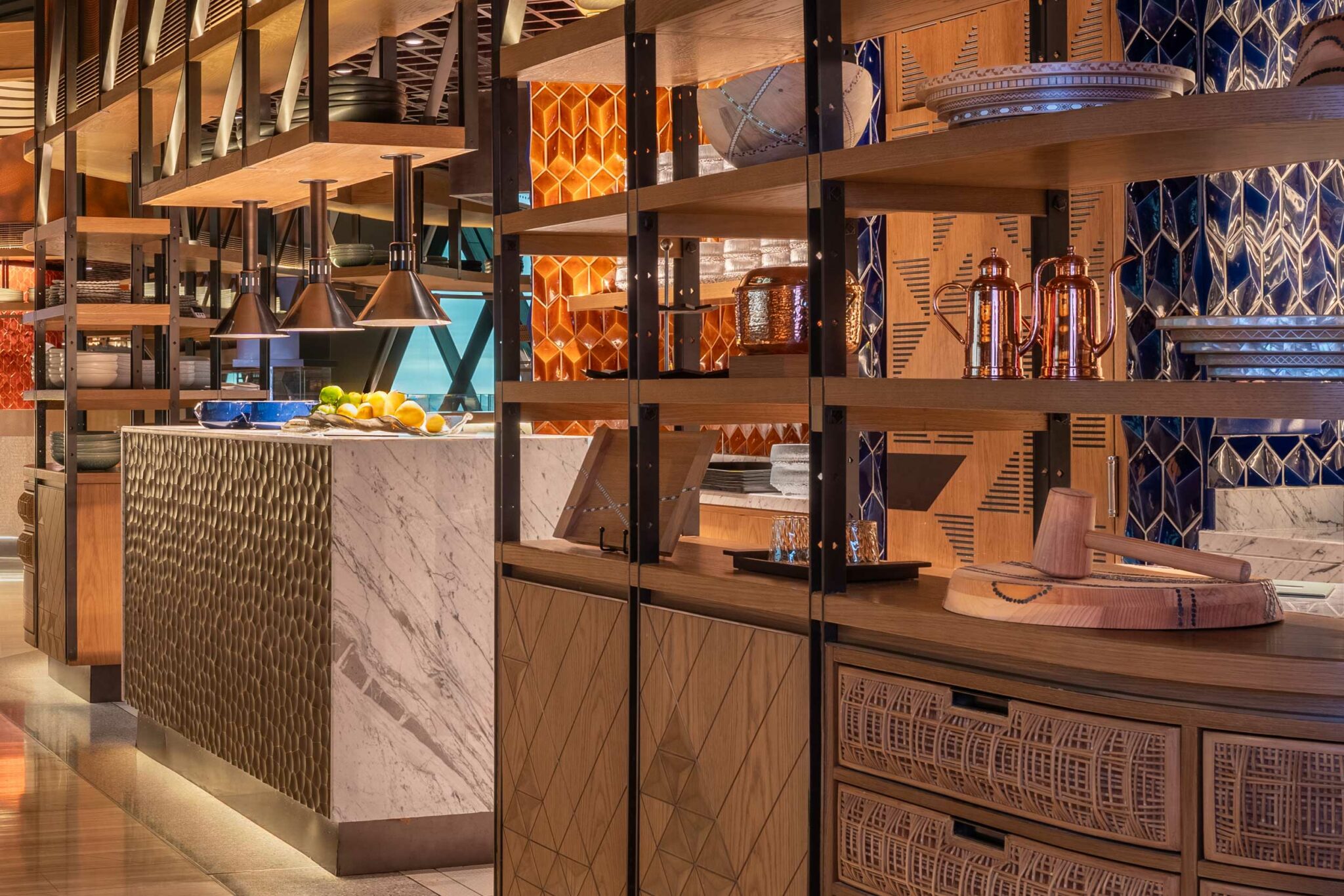 Open kitchen scene at Arrazuna, featuring marble counters, warm wood shelving, copper cookware, and geometric tilework; fresh lemons and plated ingredients sit beneath overhead heat lamps. Reminiscent of a lively Market.
