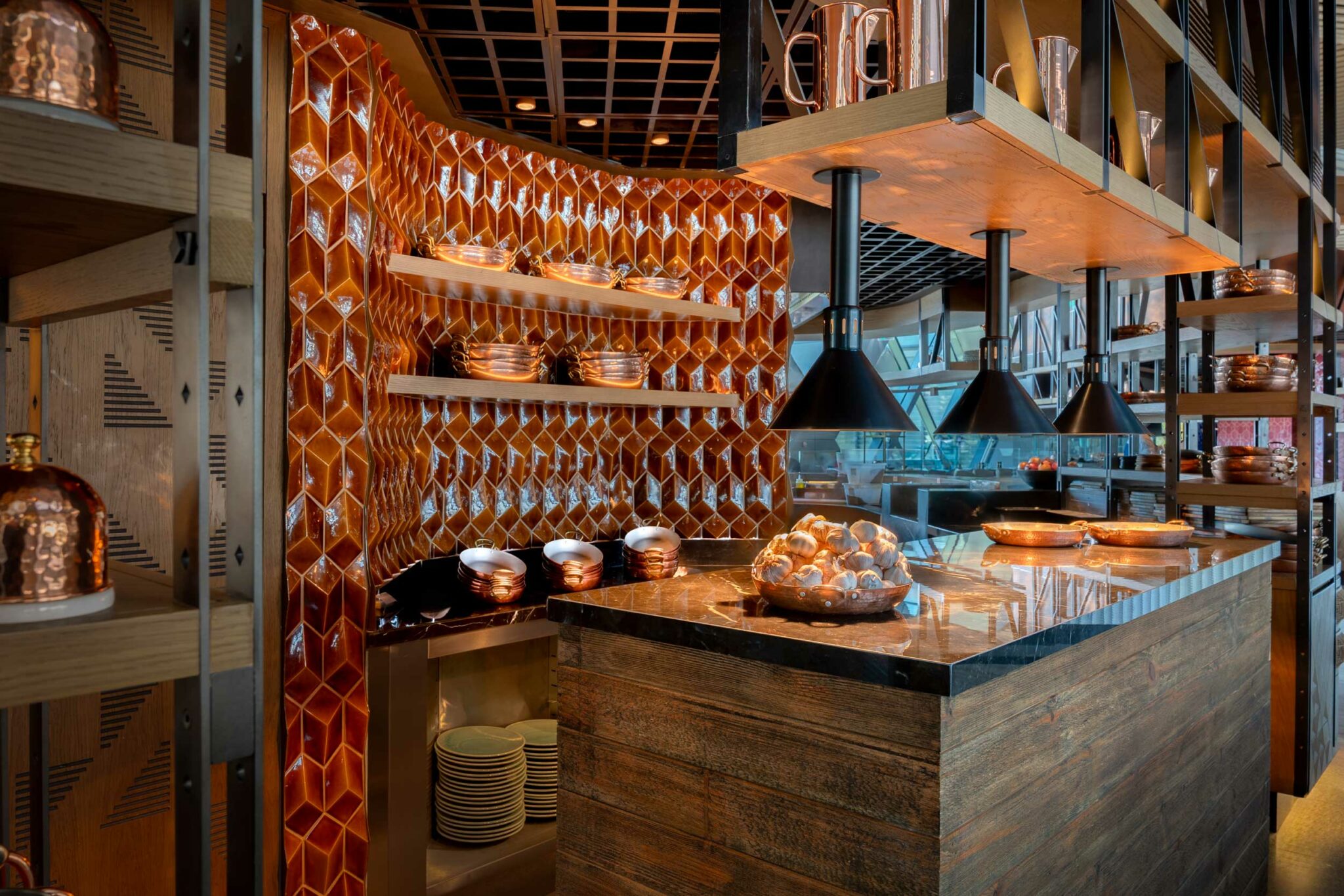 Sleek food hall interior with warm, geometric orange tiles and wooden shelves displaying copper kitchenware. The countertop glows under modern black lamps.