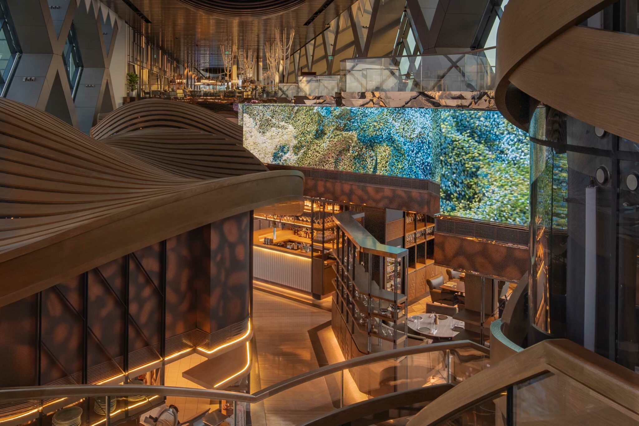 Luxurious, modern interior with wavy wooden textures and warm lighting. A large, vibrant digital art screen is mounted on the wall above seating areas.