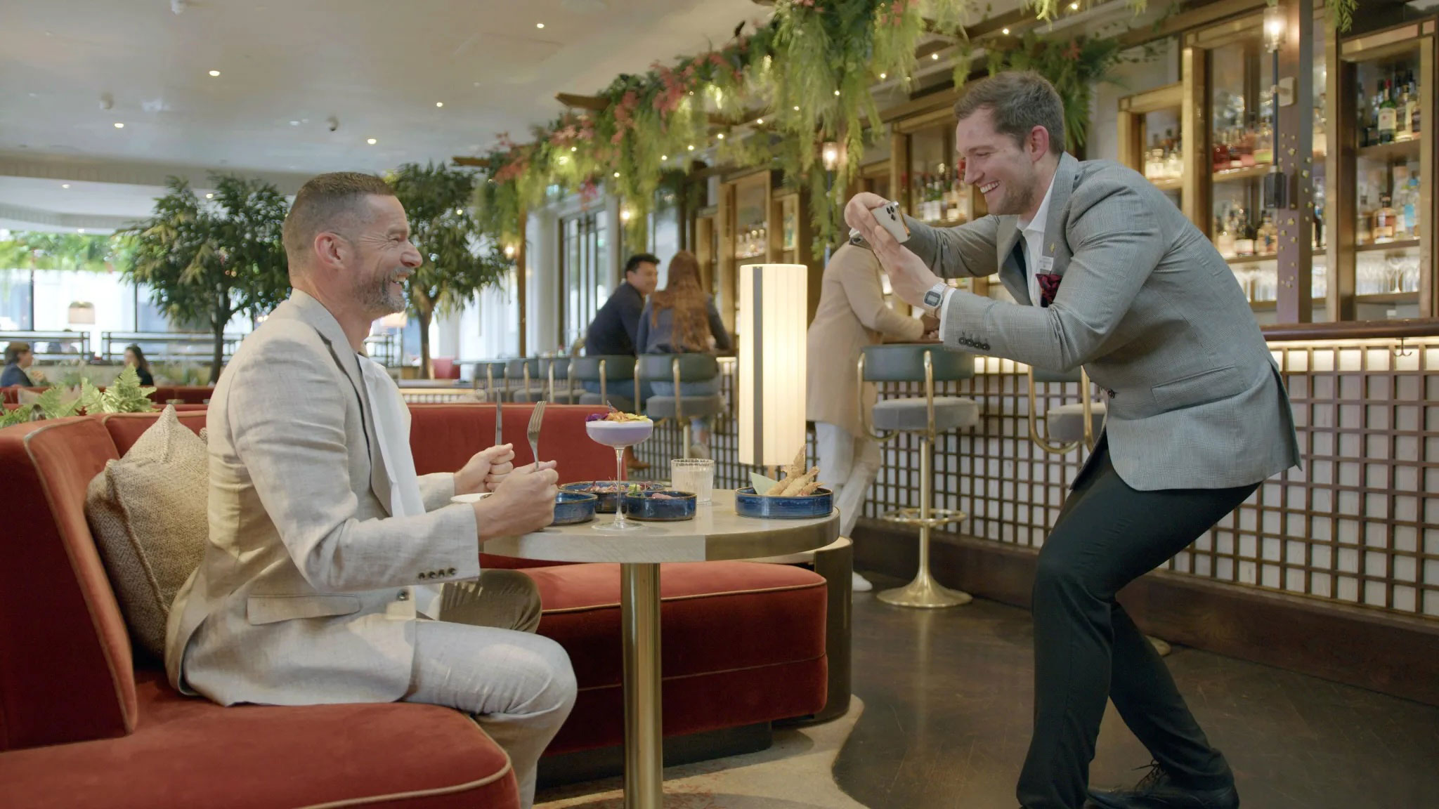 Guest being photographed by waitress in Hilton hotel. They hare a playful moment as one photographs the other’s dishes in a stylish bar lounge filled with greenery, warm lighting, and velvet seating—capturing a modern hospitality setting designed for social, photogenic dining experiences.
