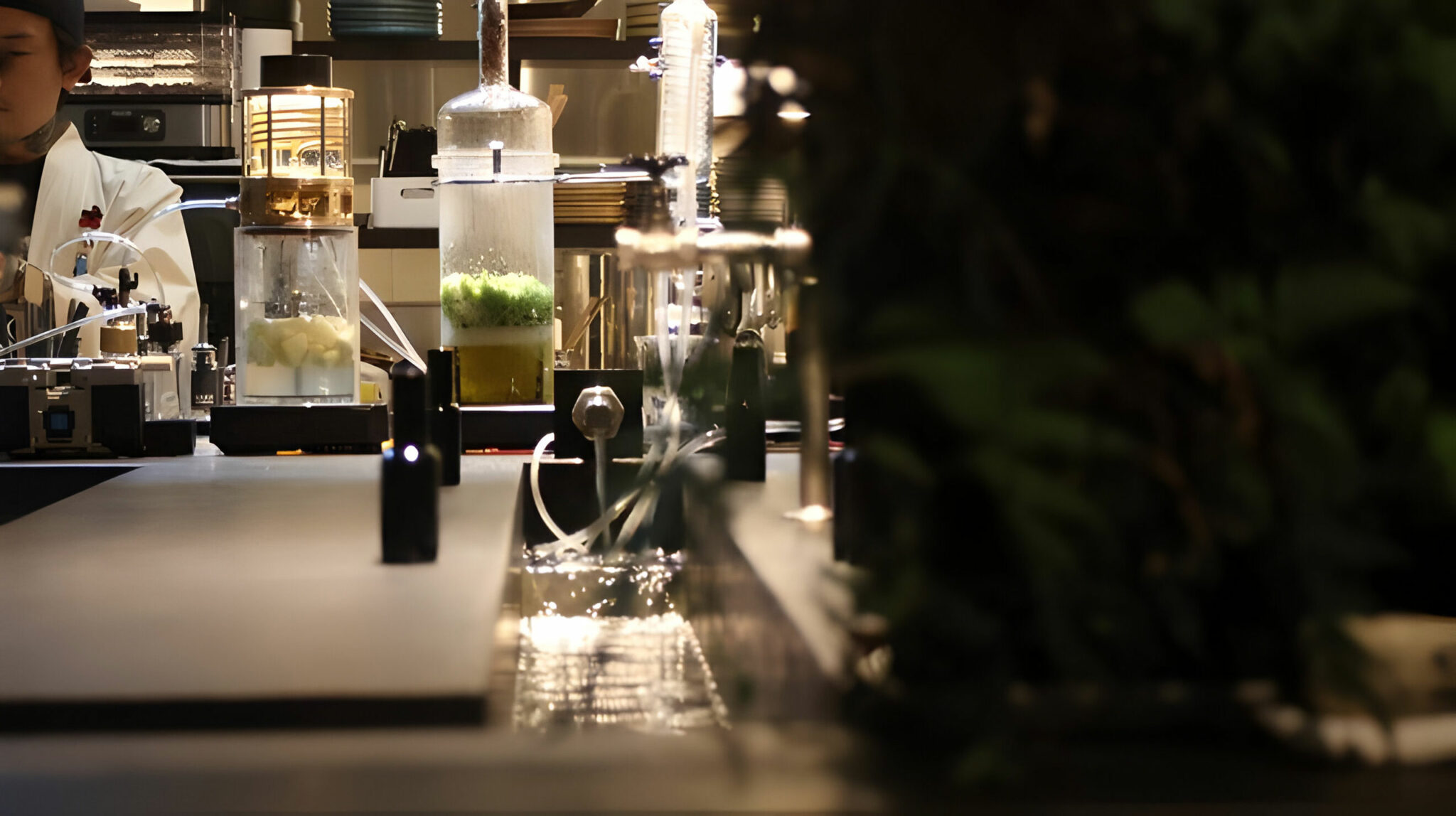 A close-up of an experimental bar or kitchen setup featuring glass infusion vessels, tubing, and illuminated apparatus, with herbs and fruits steeping inside. Located in Japan called Cocktail Stand Furek.