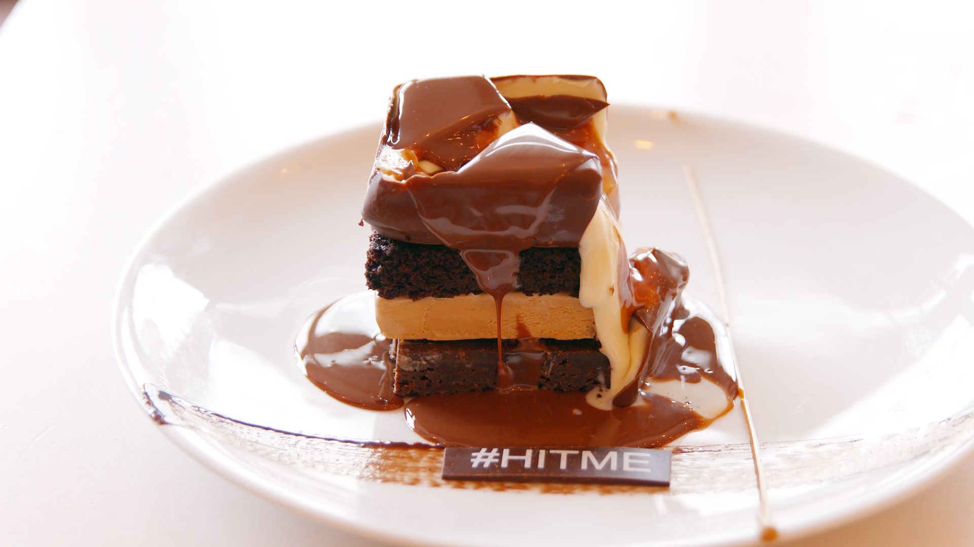 A decadent stacked dessert of brownies, caramel, and ice cream drenched in warm chocolate sauce, served on a white plate with a “#HITME” chocolate tag—capturing a playful, indulgent food presentation designed for dramatic, guest-interactive moments. Catch LA.