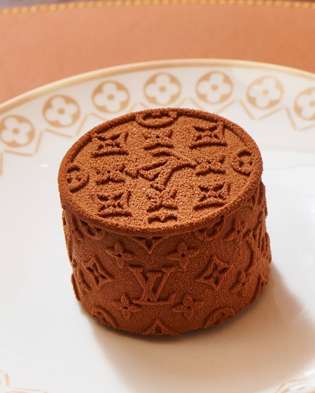 A chocolate mousse dessert embossed with a louis vuitton monogram pattern, presented on an elegant patterned plate—showcasing elevated, brand-driven culinary design that blends fashion aesthetics with fine dining craftsmanship.