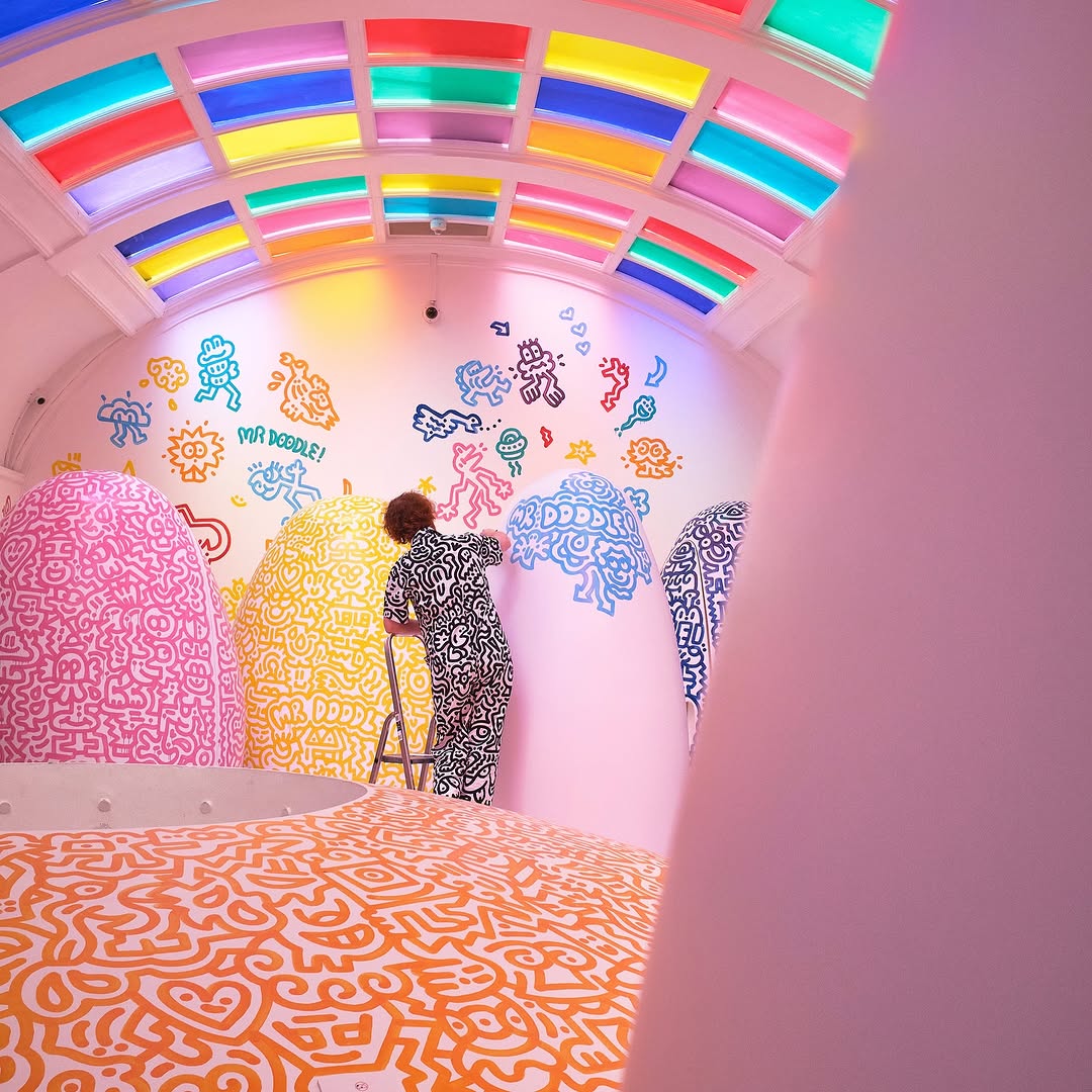 An artist in a patterned jumpsuit paints colourful doodle-style illustrations across a curved, pastel room filled with oversized sculptural forms, under a ceiling of rainbow glass panels—showcasing immersive, playful spatial design and creative brand storytelling.