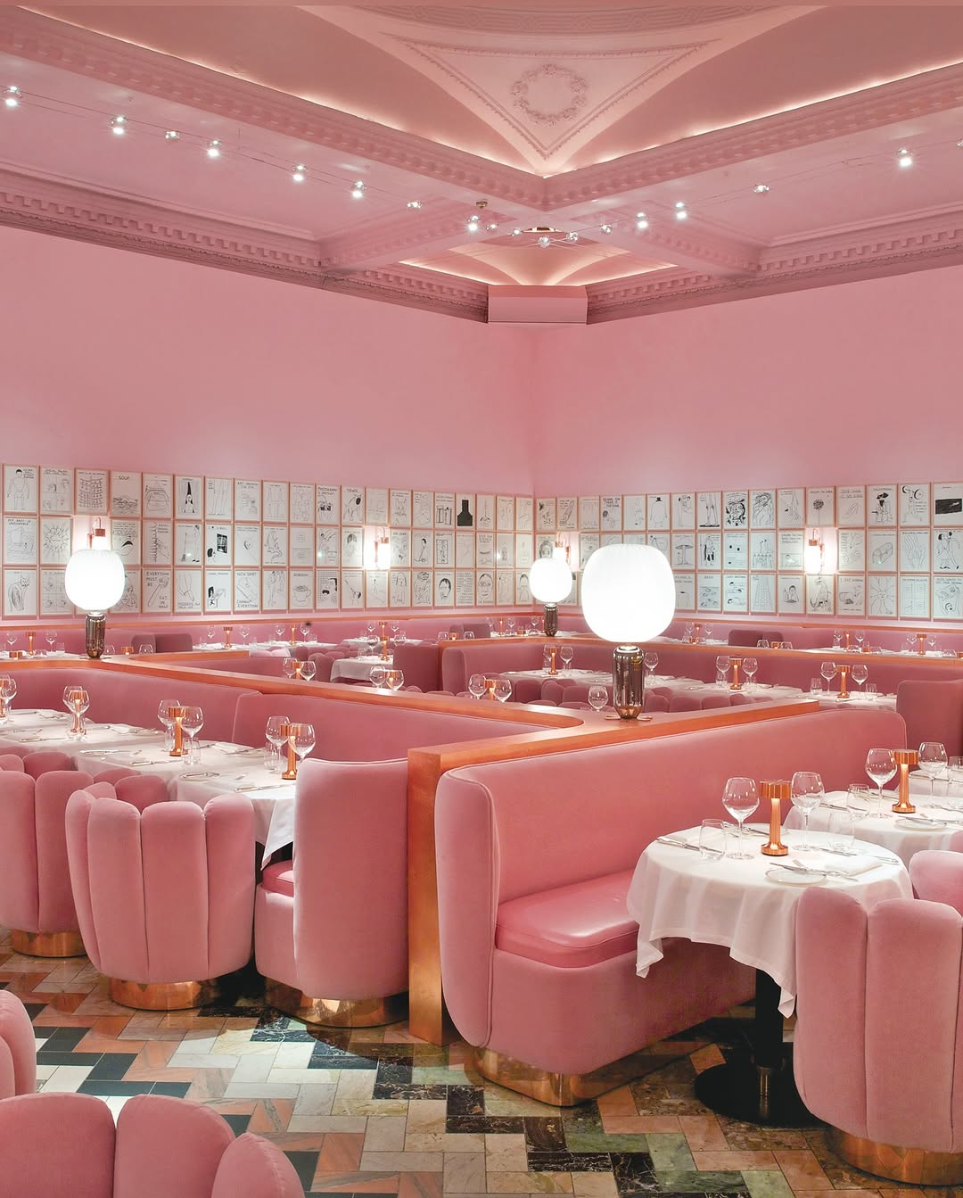A playful, design-driven dining room in London called Sketch fully immersed in blush pink, with sculpted velvet banquettes, round white-clothed tables, oversized globe lamps, and walls lined with framed illustrations—showcasing bold, immersive hospitality interior design with a strong artistic identity.