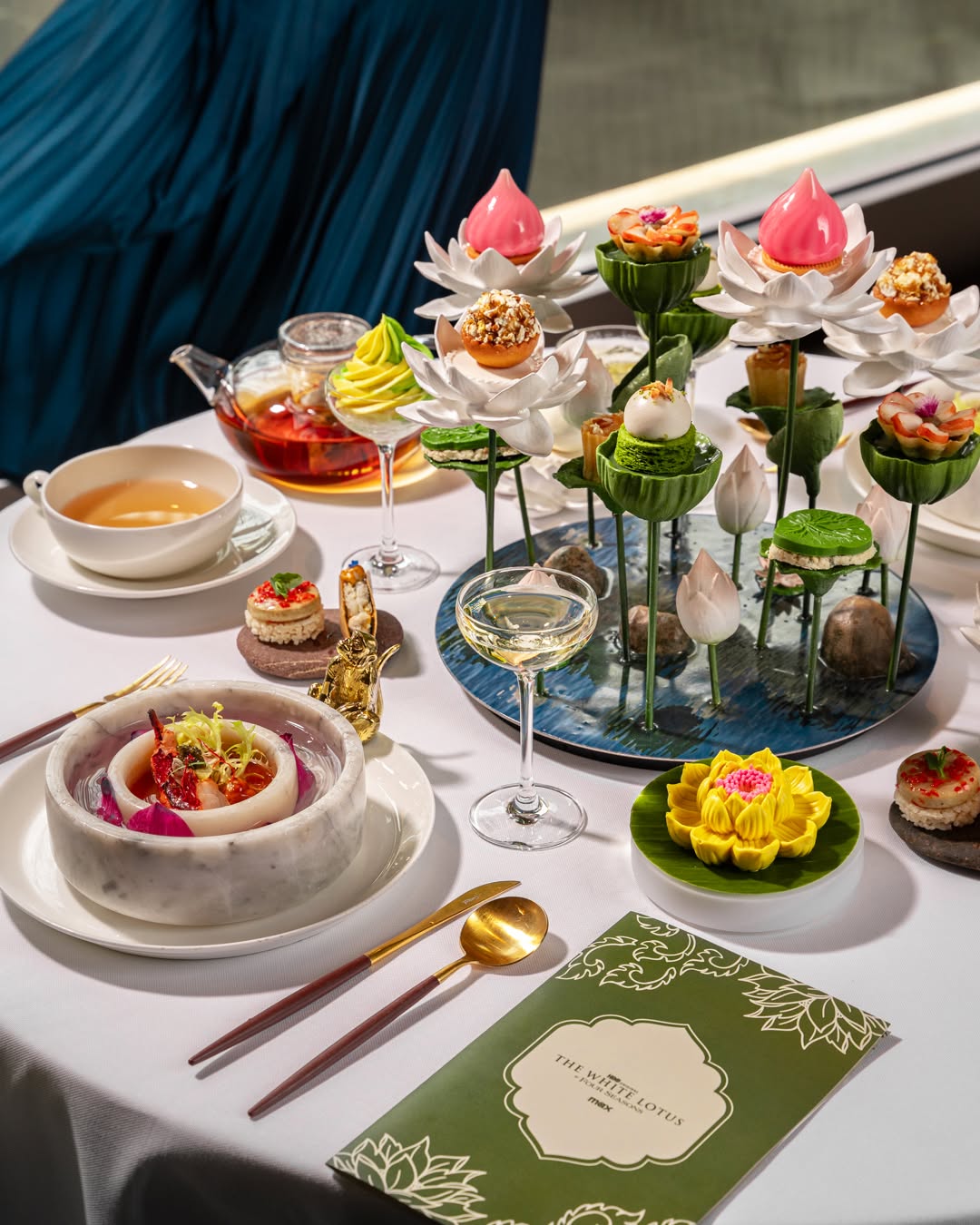 A Four Seasons HBO White Lotus collabration afternoon tea presentation inspired by lotus flowers, featuring intricate pastries displayed on sculptural lotus stands, a marble bowl dish, fine tea service, and floral details—illustrating theatrical, storytelling-driven hospitality design for luxury dining experiences.
