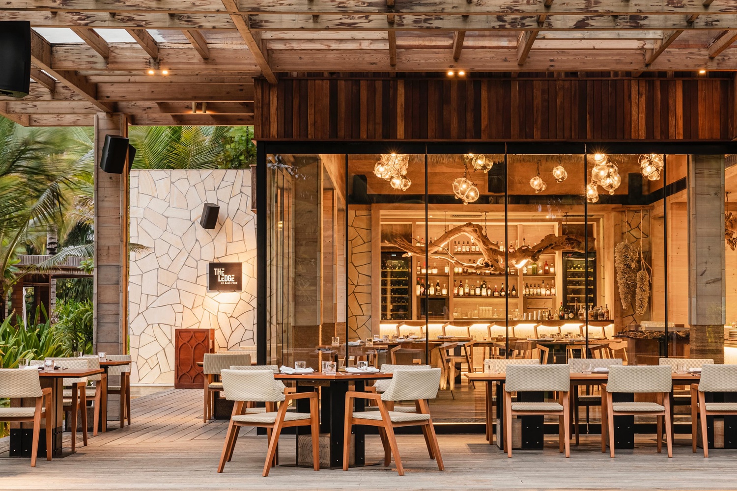 Open-air dining terrace at The Ledge by Dave Pynt, Waldorf Astoria Maldives, showcasing Social F+B’s interior design with warm timber architecture, sculptural lighting, floor-to-ceiling glazing, and a central open kitchen overlooking tropical surroundings.