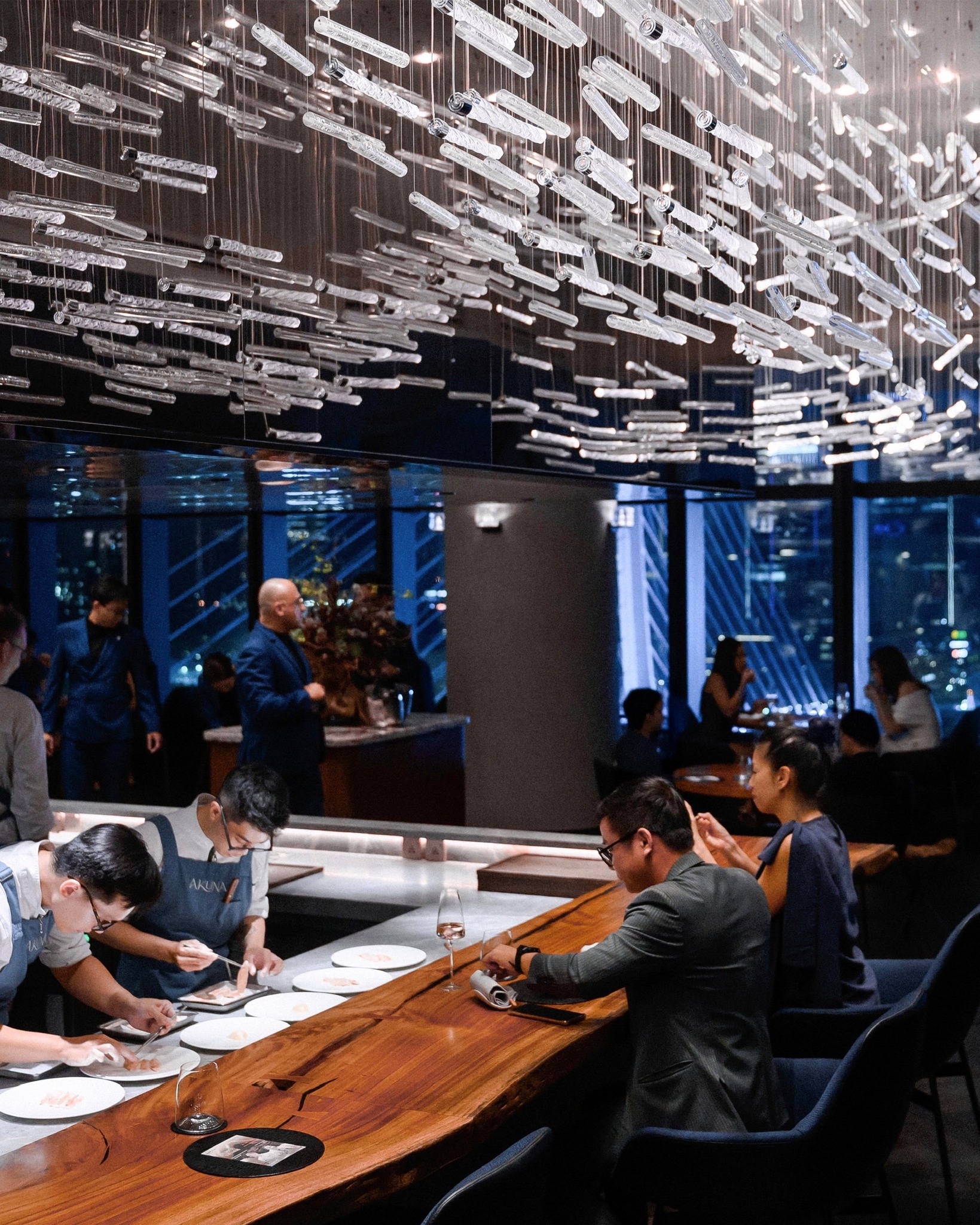 Interior of Akuna Restaurant at Le Méridien HCMC City, featuring a chef’s counter dining experience beneath a sculptural installation of suspended glass rods, with guests seated along a live preparation bar