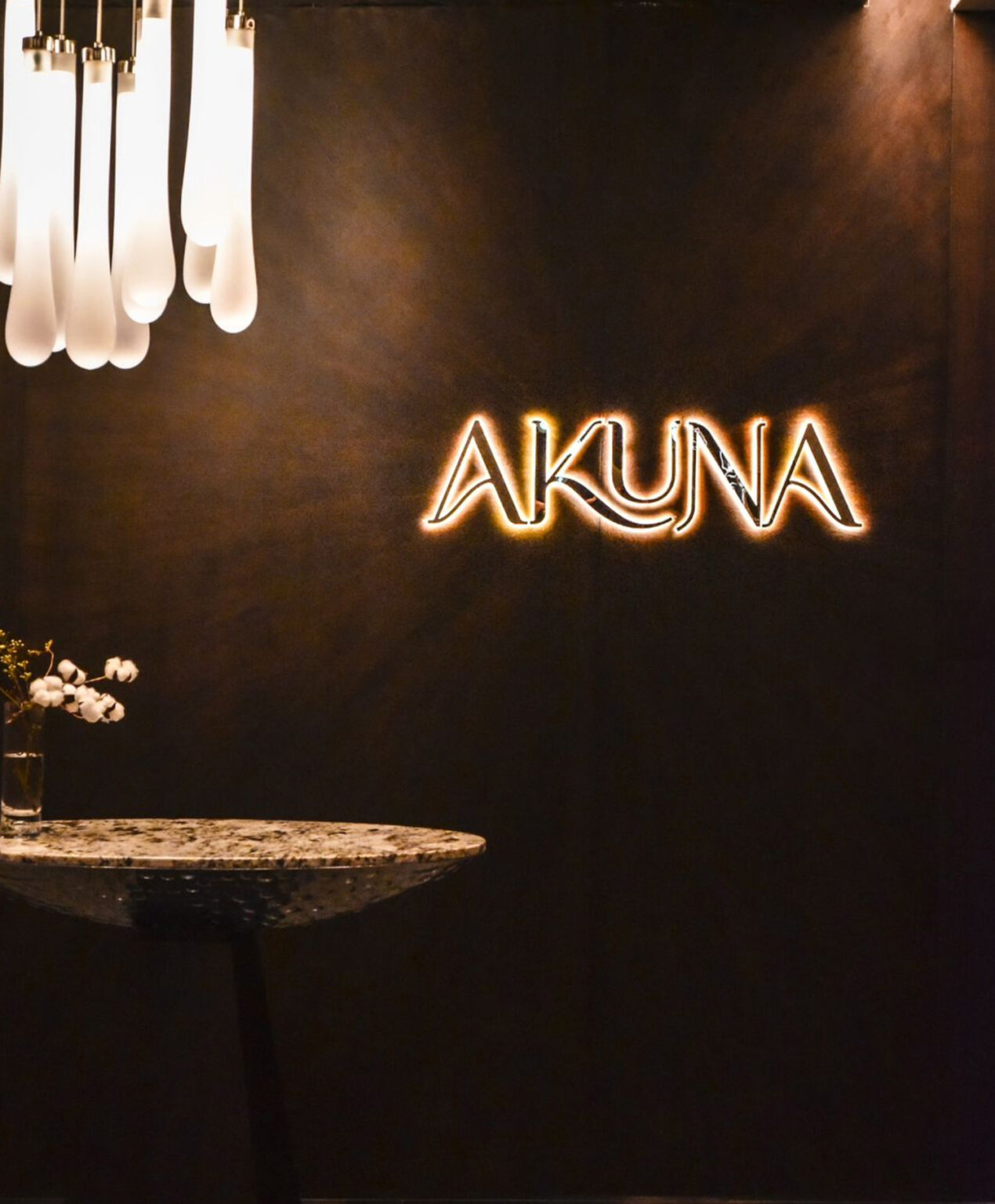 Illuminated Akuna Restaurant signage mounted on a dark textured wall, paired with sculptural pendant lighting and a stone table, reflecting the refined brand identity designed by Social F+B at Le Méridien HCMC City.