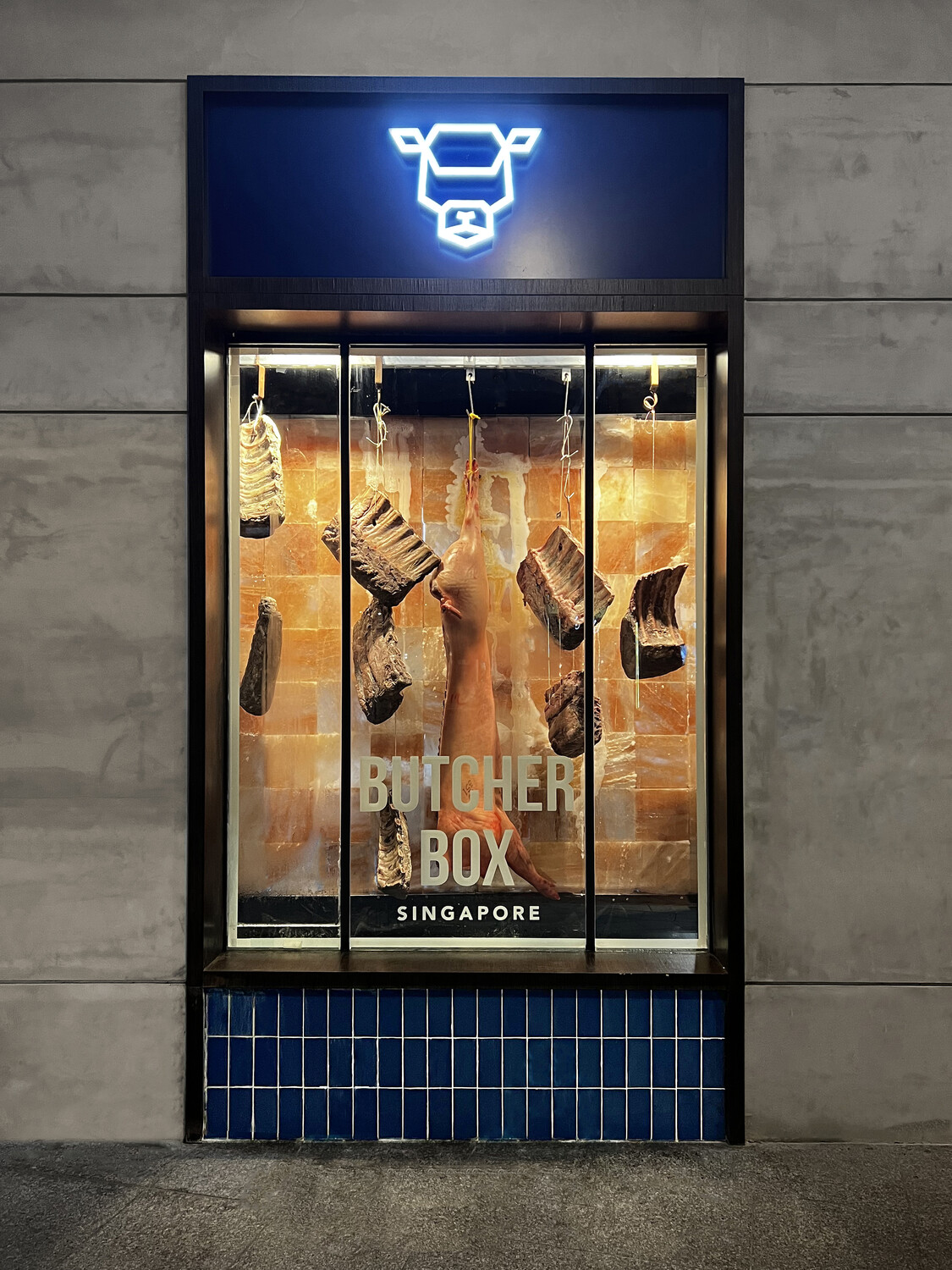 Exterior window display of Butcher Box Singapore featuring illuminated cow icon, Himalayan salt brick backdrop, and hanging cuts of meat—showcasing Social F+B’s premium butcher retail design and brand experience.