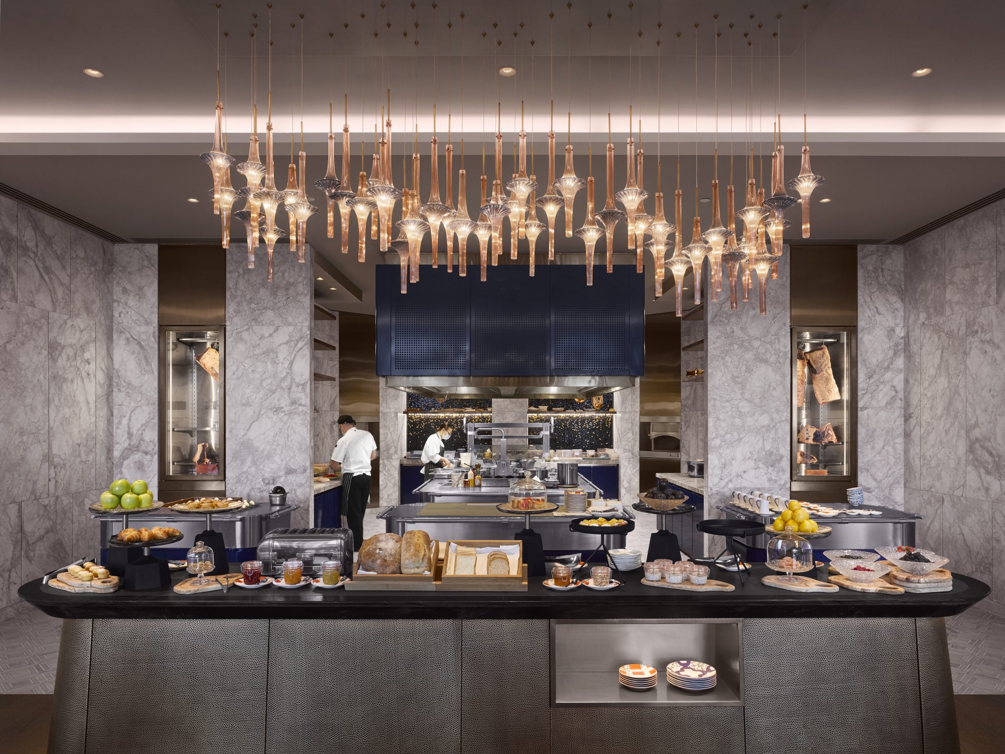 Interior of La Veranda, the all-day dining restaurant at One&Only Portonovi, Montenegro, featuring an open kitchen framed by marble columns, sculptural glass lighting, and a central buffet counter showcasing artisanal breads and produce within a refined, contemporary hospitality setting.